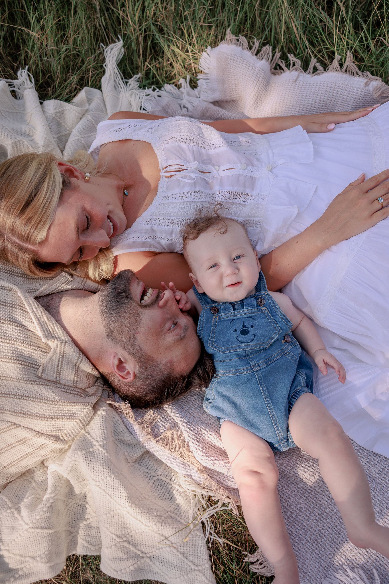 Family with baby lying on blanket in grass, parents smiling at infant in denim overalls during outdoor picnic in South Cerney, Cotswolds.