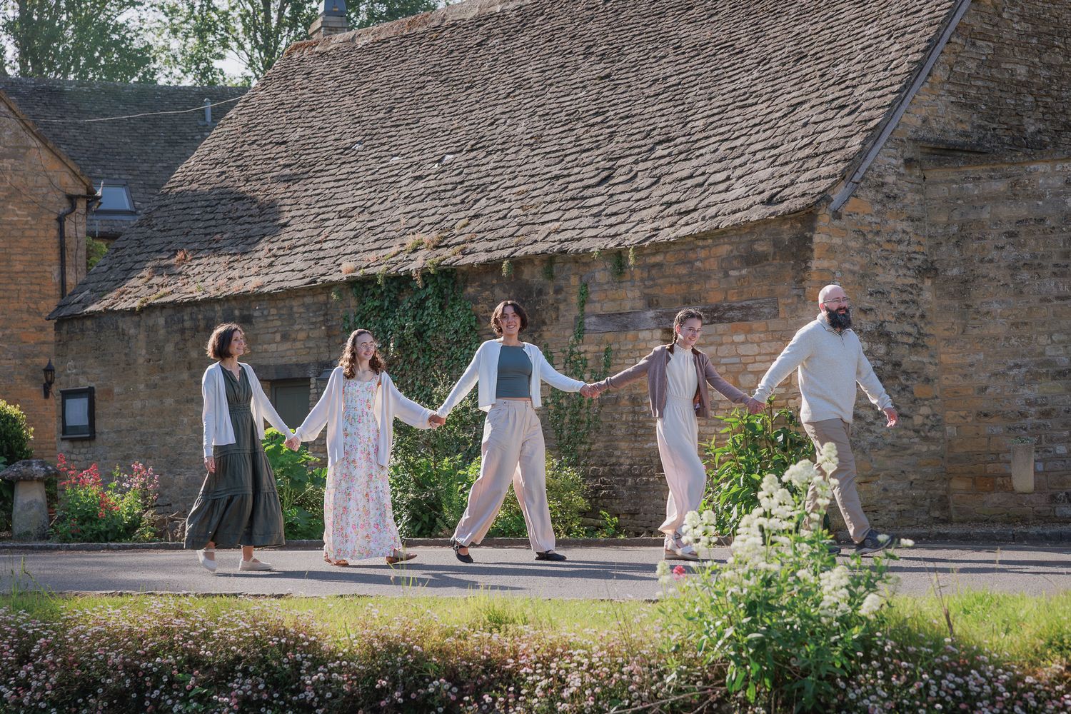 Five people holding hands walking together outside a rustic stone cottage in a sunny English countryside village.