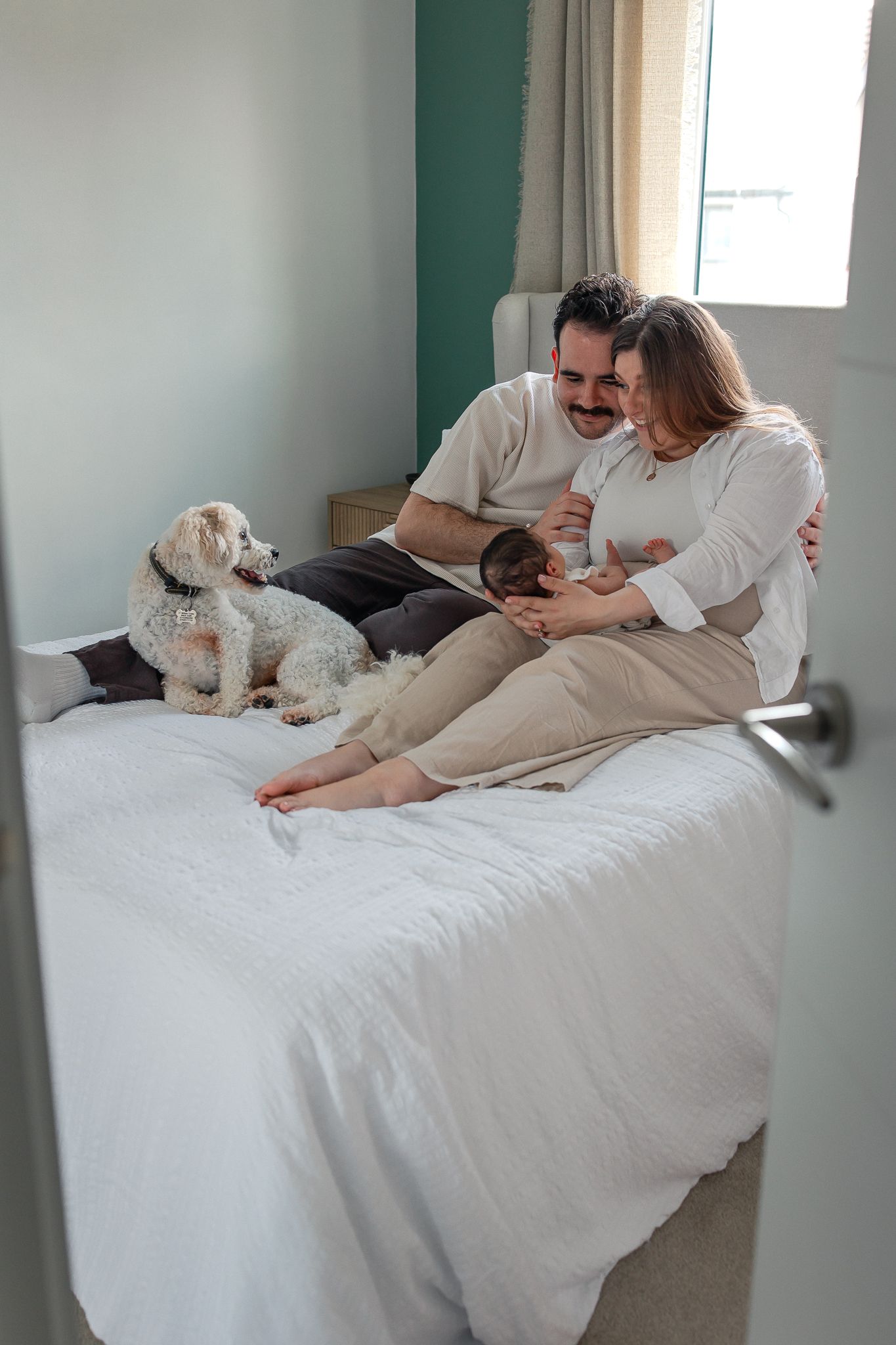A couple sits on a bed with their newborn baby and fluffy white dog in a cozy bedroom in Corsham.