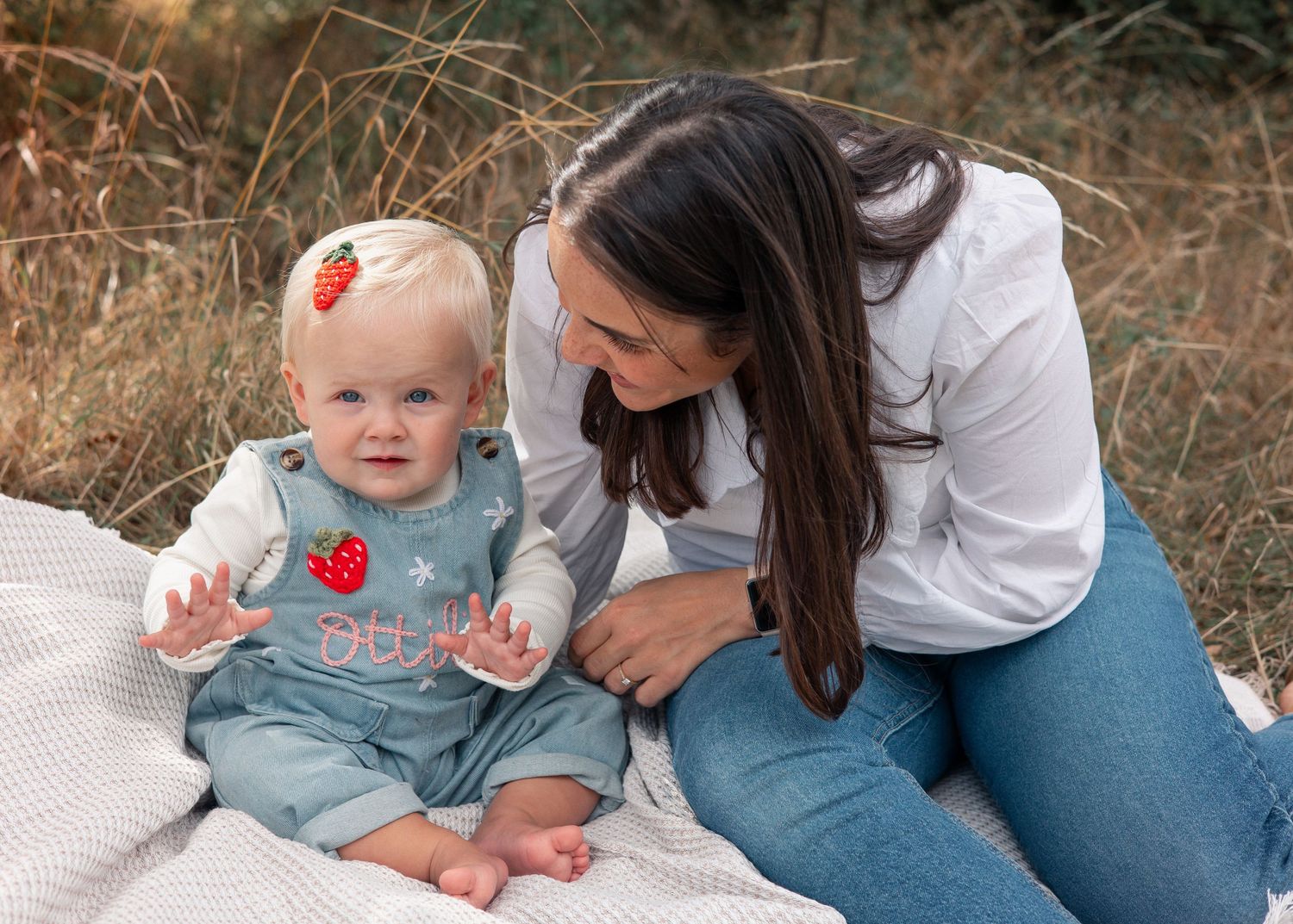 A mother smiles lovingly at her baby girl in a strawberry outfit on a blanket in a golden field.
