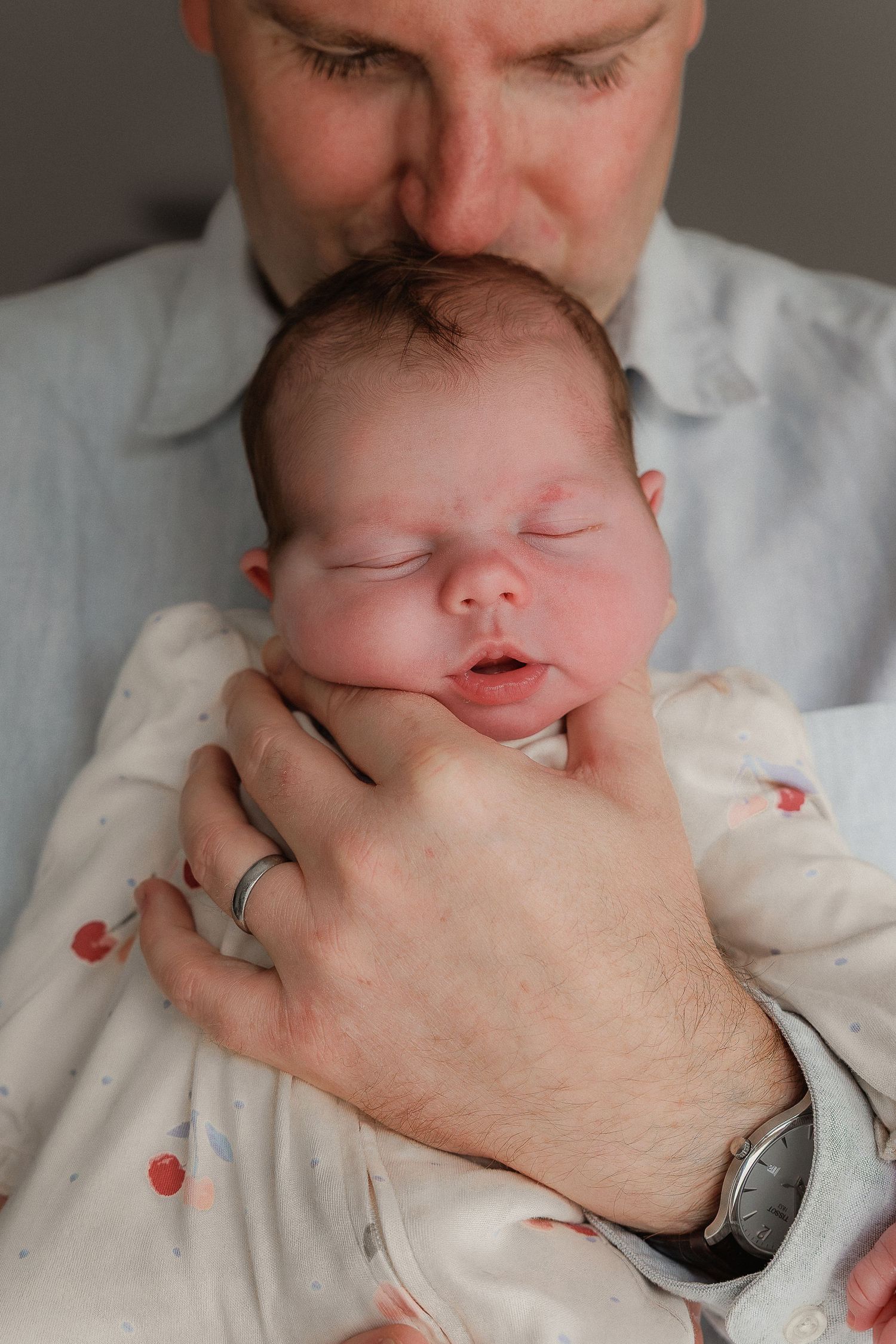 Newborn Photographer takes photo of father gently kissing his sleeping newborn baby in Cirencester Cotswolds.