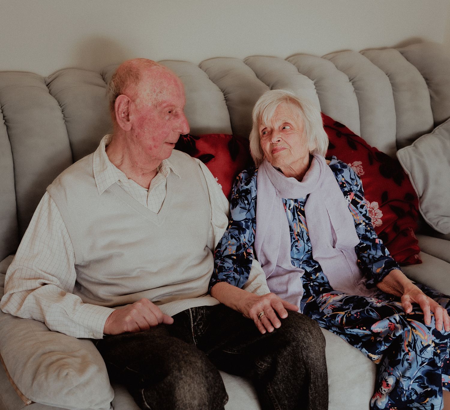 Elderly couple sitting together on a beige sofa, sharing a warm moment and looking at each other fondly in Chipping Campden.