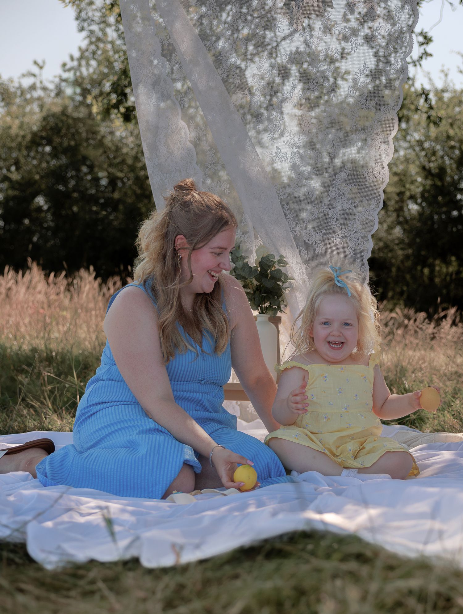 A woman in a blue dress and toddler girl in yellow sit laughing on a white blanket outdoors on a sunny day in Bibury, Cotswolds.