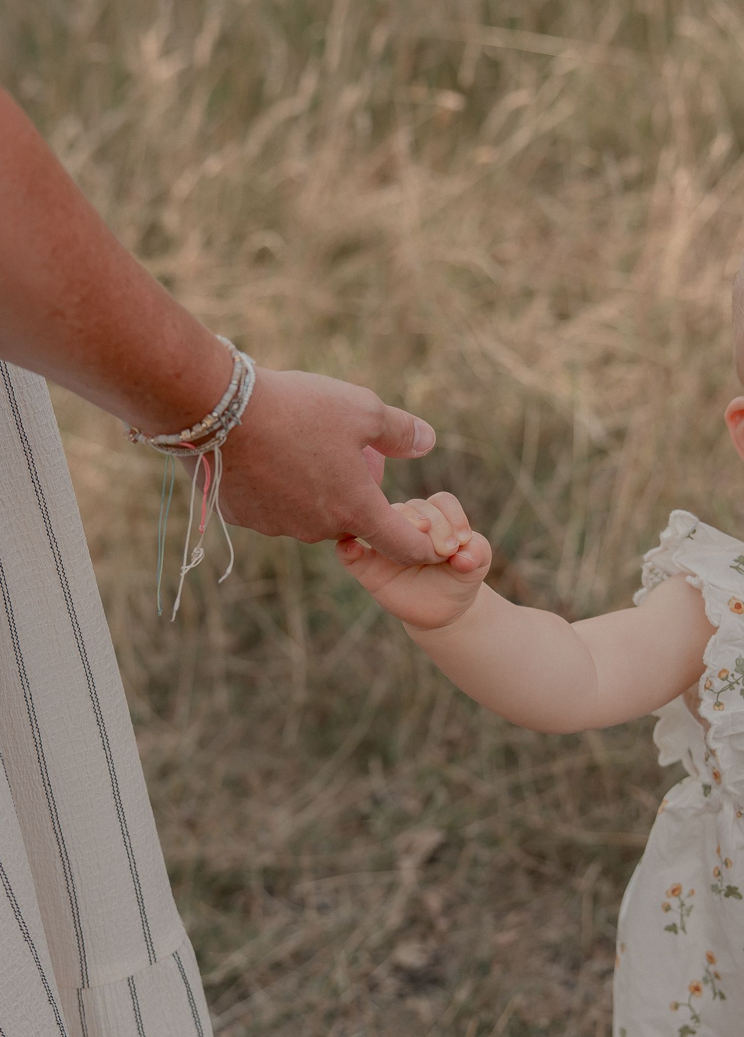 Adult hand with bracelets gently holding a baby's tiny hand outdoors in a golden grass field.