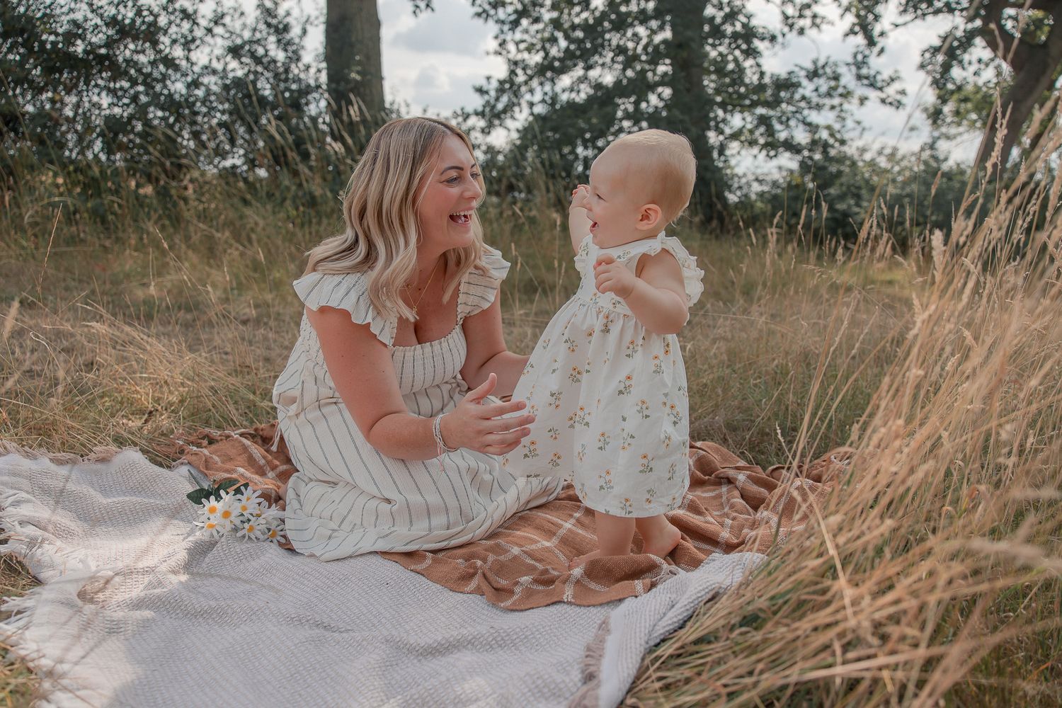 A mother and baby girl sit on a blanket in a field, sharing a joyful moment in matching floral outfits.