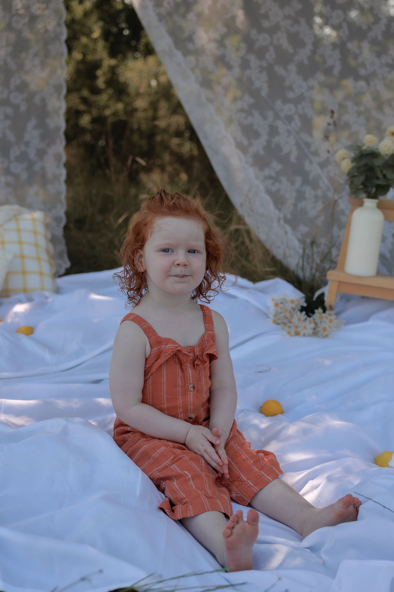 A young redhead girl in a rust-colored romper sits on a white blanket near lace curtains and lemons in Henley-on-Thames, Oxfordshire.