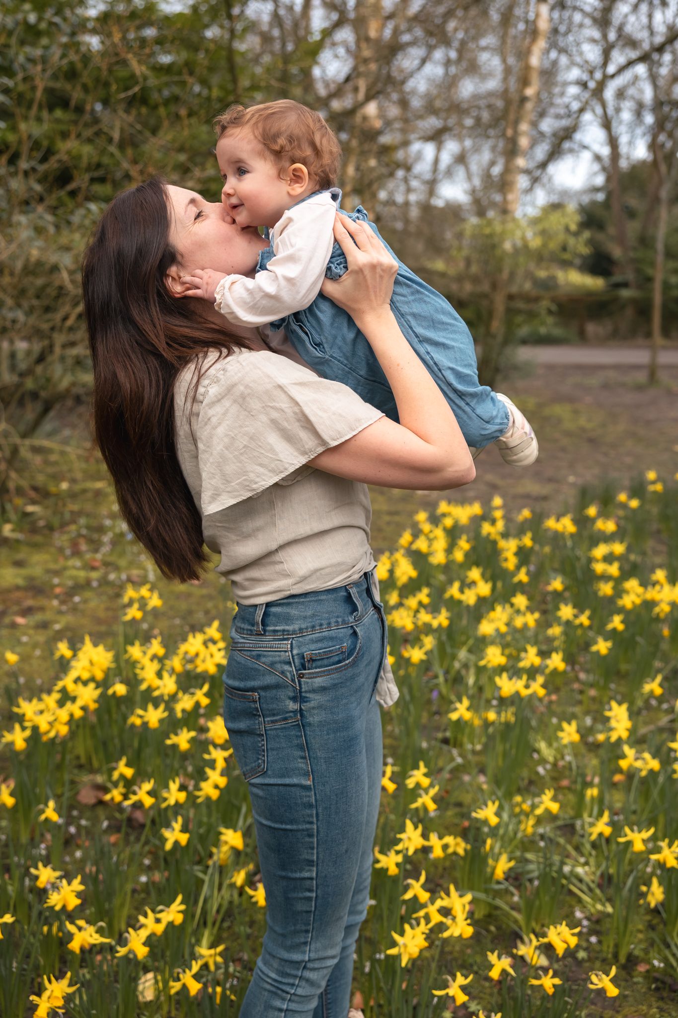 A mother lifts and kisses her baby among yellow daffodils in a spring park setting in South Cerney, Cotswolds.