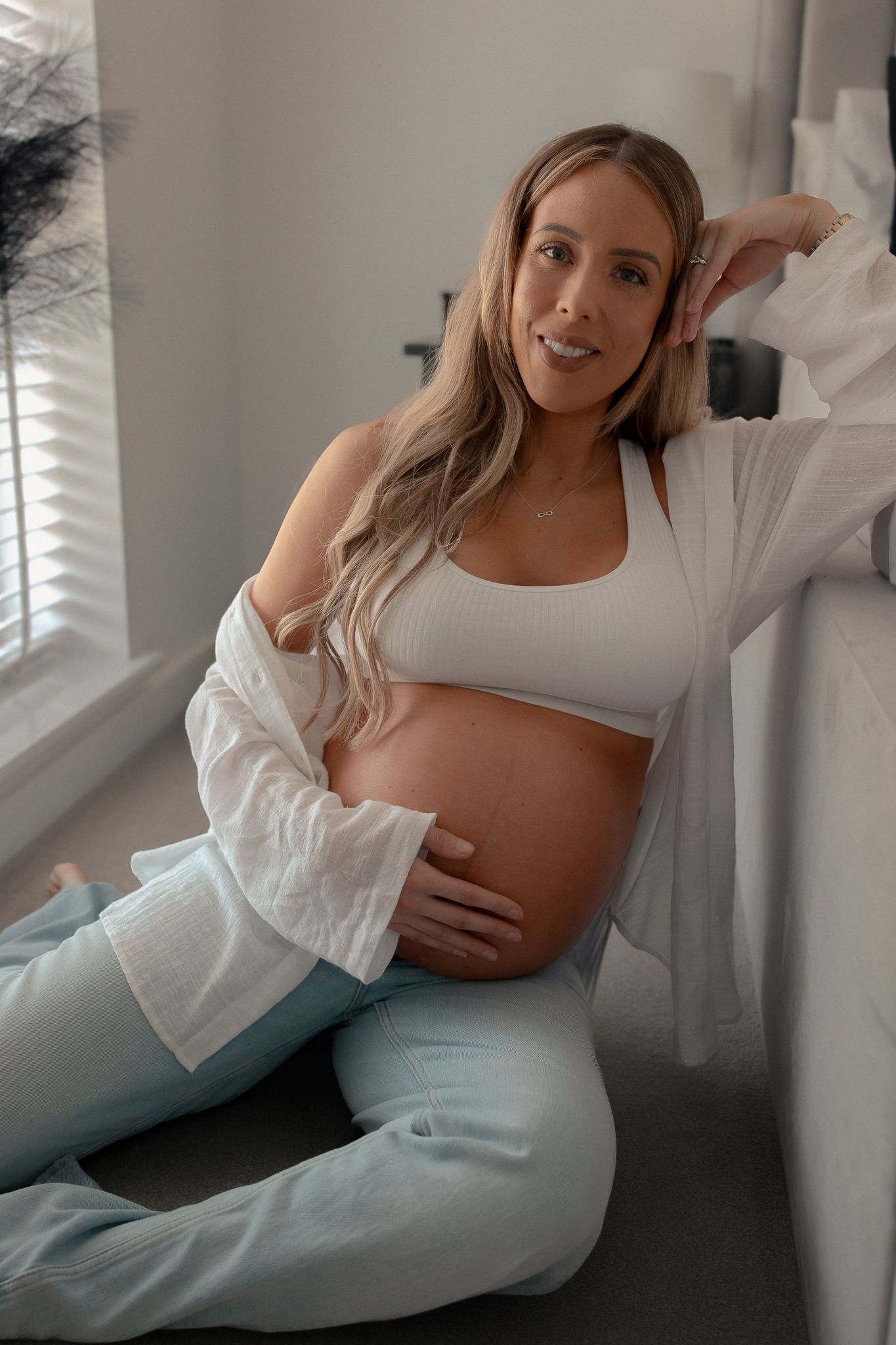 Smiling pregnant woman in white crop top and open shirt cradling her baby bump while sitting against a wall at Lakes by Yoo.