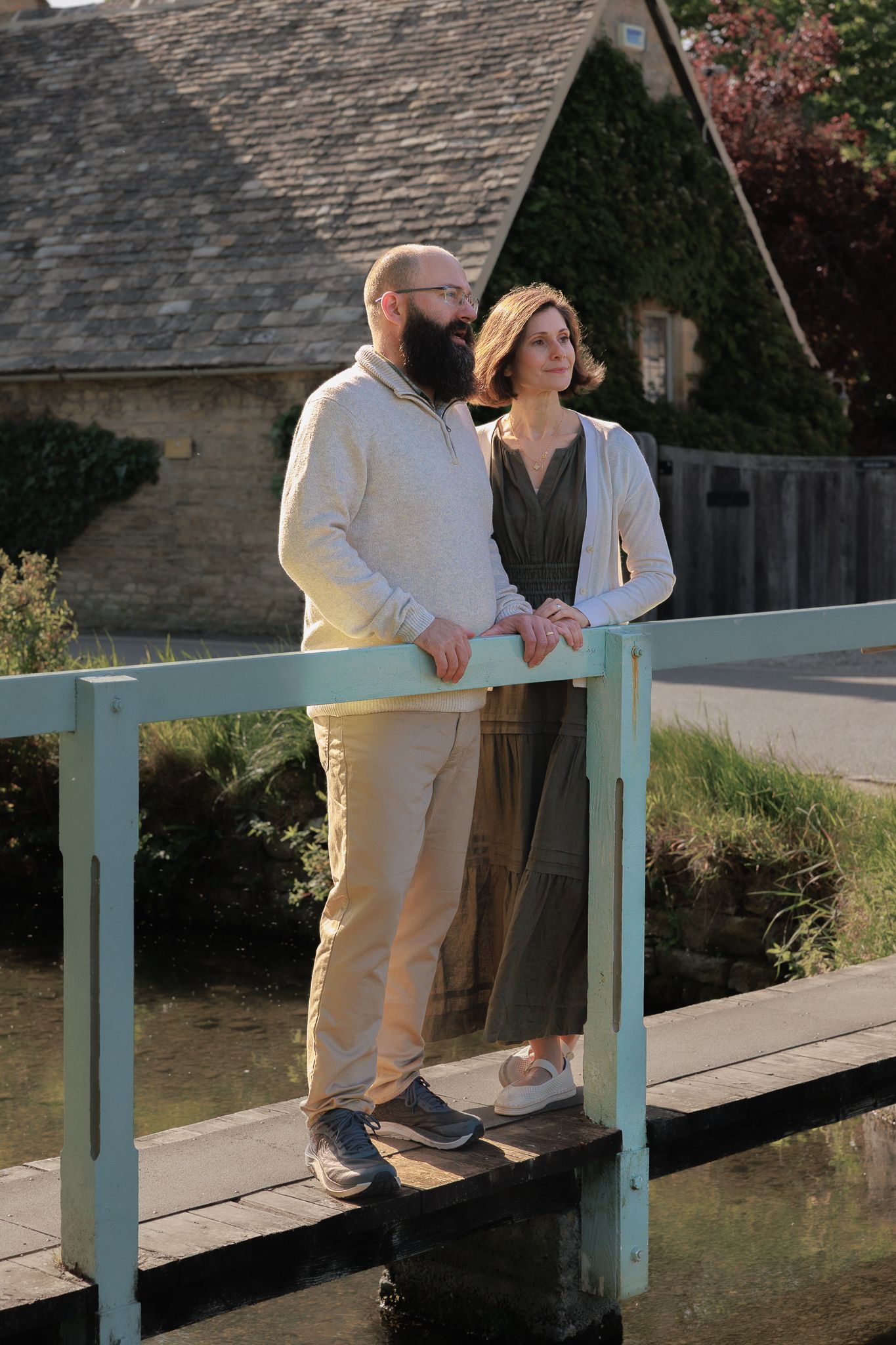 Couple standing together on a small bridge overlooking a peaceful river beside a charming stone cottage.