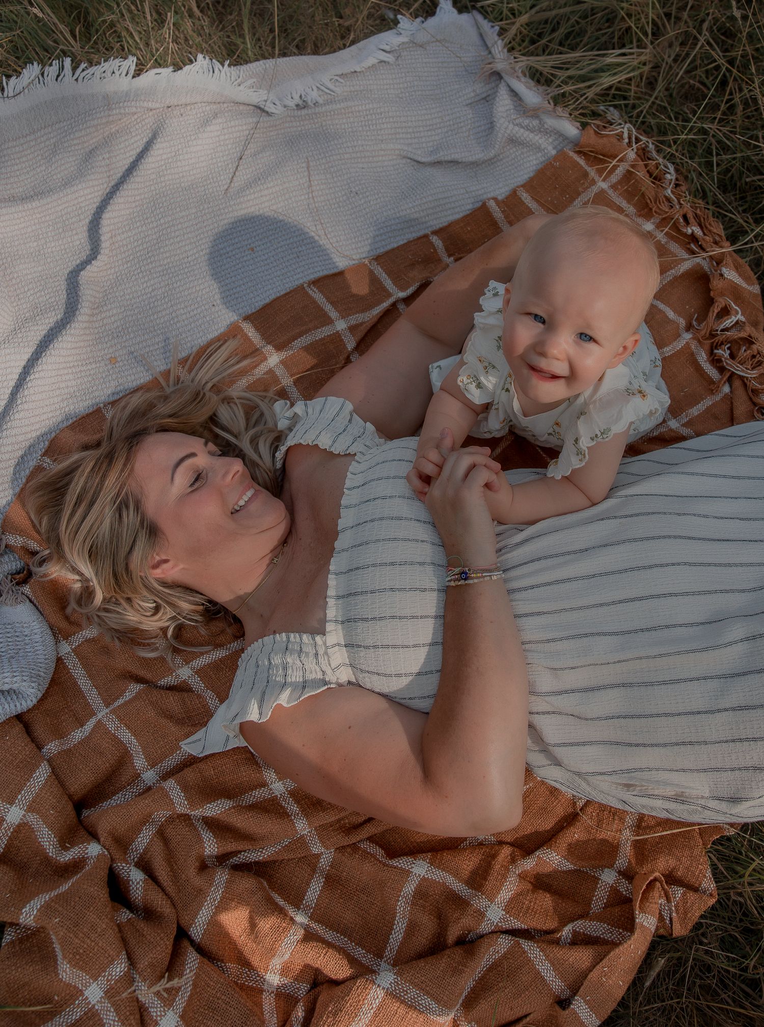 Mother and baby lying on a plaid blanket outdoors, smiling and holding hands.