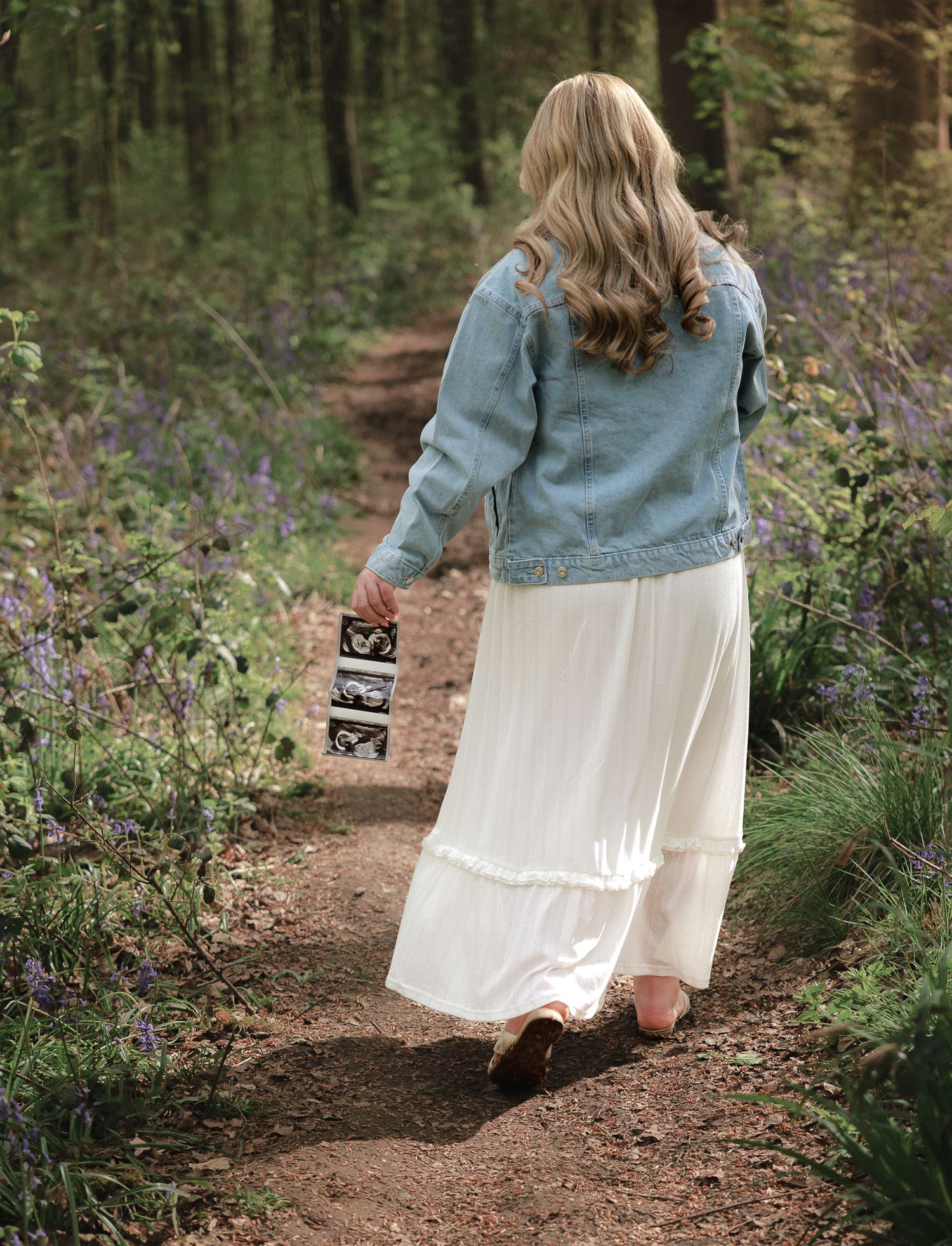 Pregnant woman walking forest path holding ultrasound photos, wearing white maxi skirt and denim jacket.