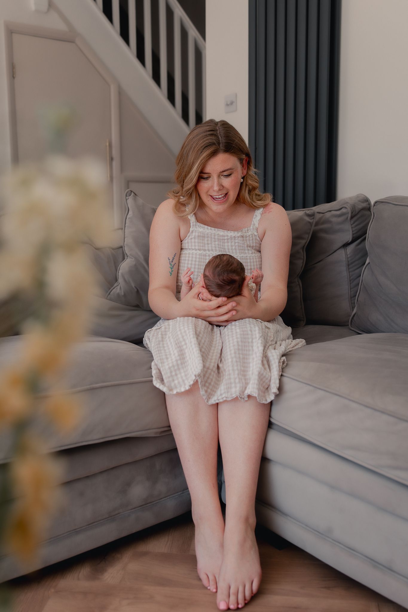 A smiling mother sits barefoot on a gray sofa, tenderly cradling her newborn baby in a bright home setting for a Newborn Photoshoot.