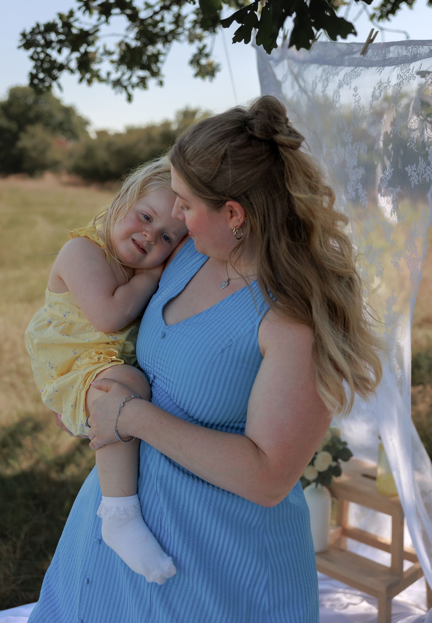 A woman in a blue dress tenderly holds a toddler in a yellow outfit outdoors in a sunny field in Bibury, Cotswolds.