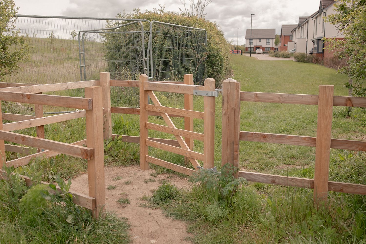 Wooden five-bar gate and fence on a rural footpath leading to a grassy field with houses in the background in Castle Eaton.