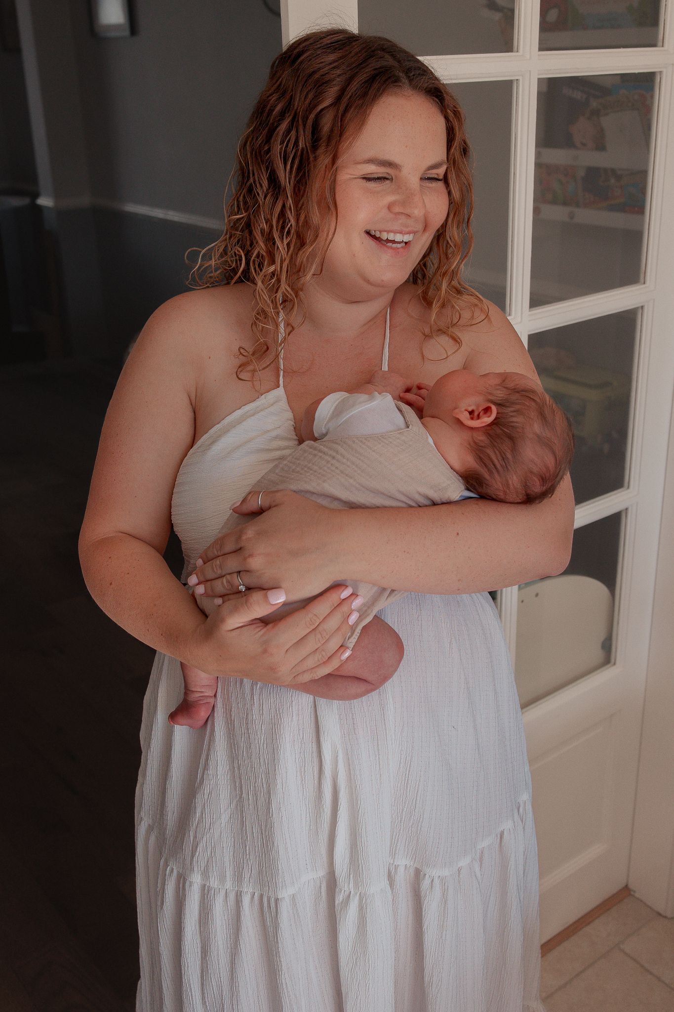 Smiling mother in white dress tenderly holding swaddled newborn baby indoors in Clevedon, North Somerset.