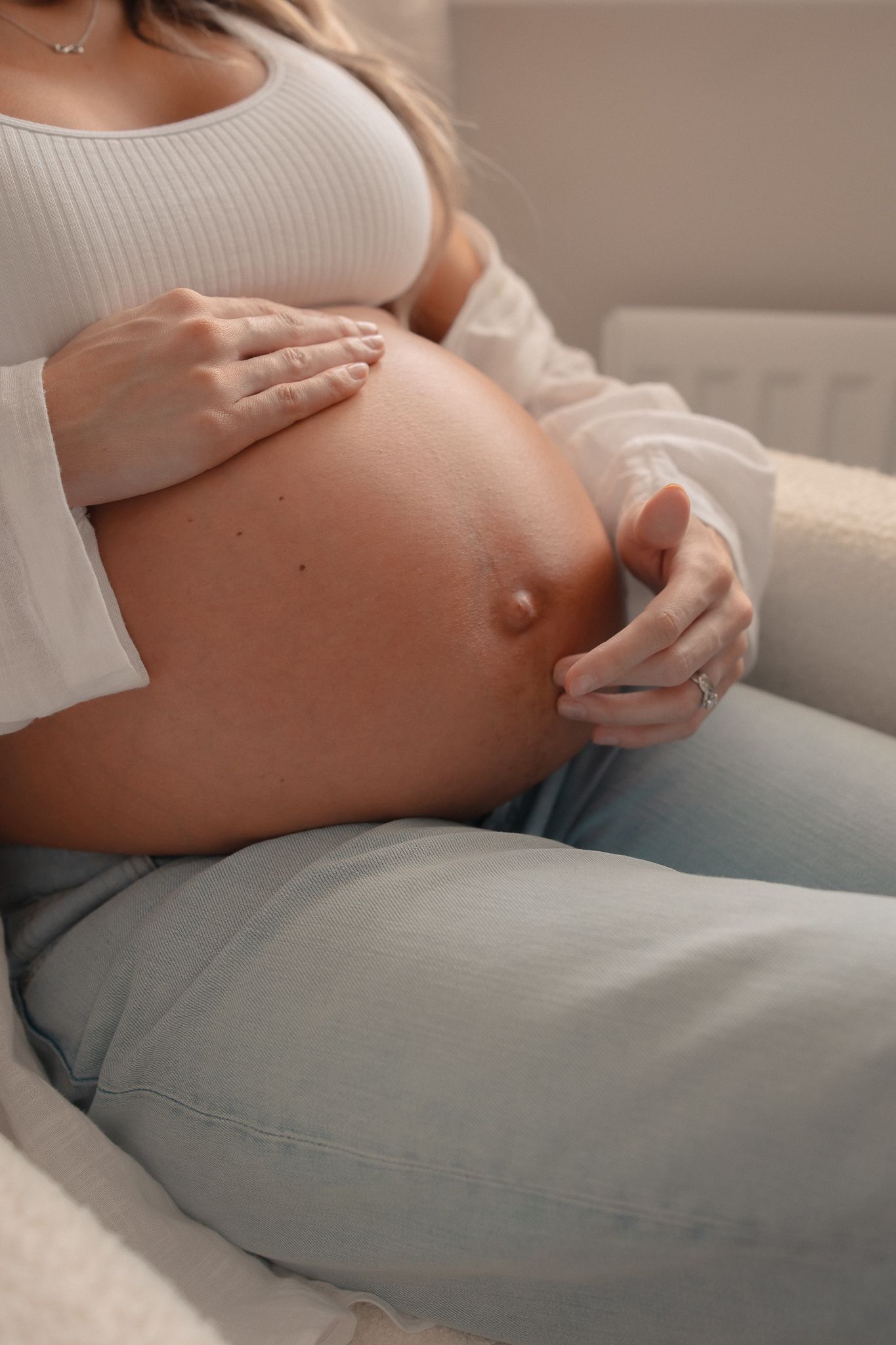 Pregnant woman in white crop top and jeans gently cradling her bare baby bump while sitting comfortably in Lechlade.