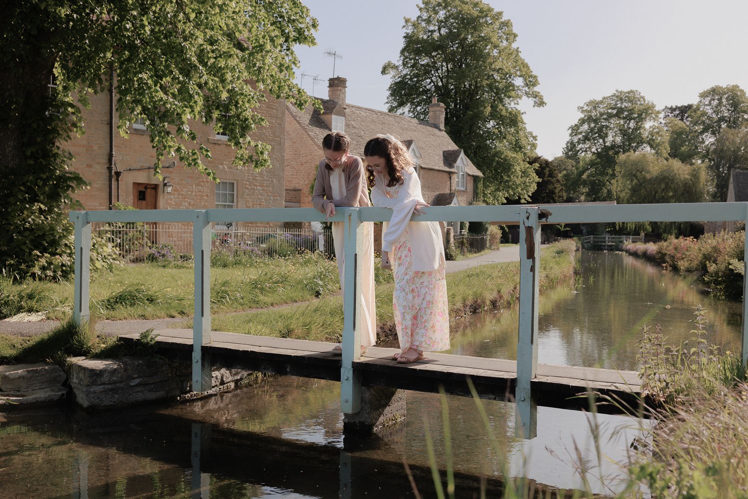 Romantic couple standing on a rustic bridge over a peaceful stream near a charming countryside home.