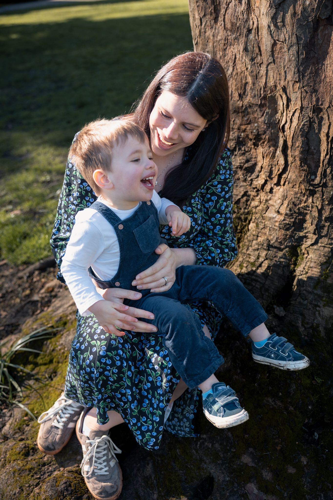 A mother and young toddler sitting together at the base of a tree in a sunny park, laughing and sharing a joyful moment in North Cerney, Cot