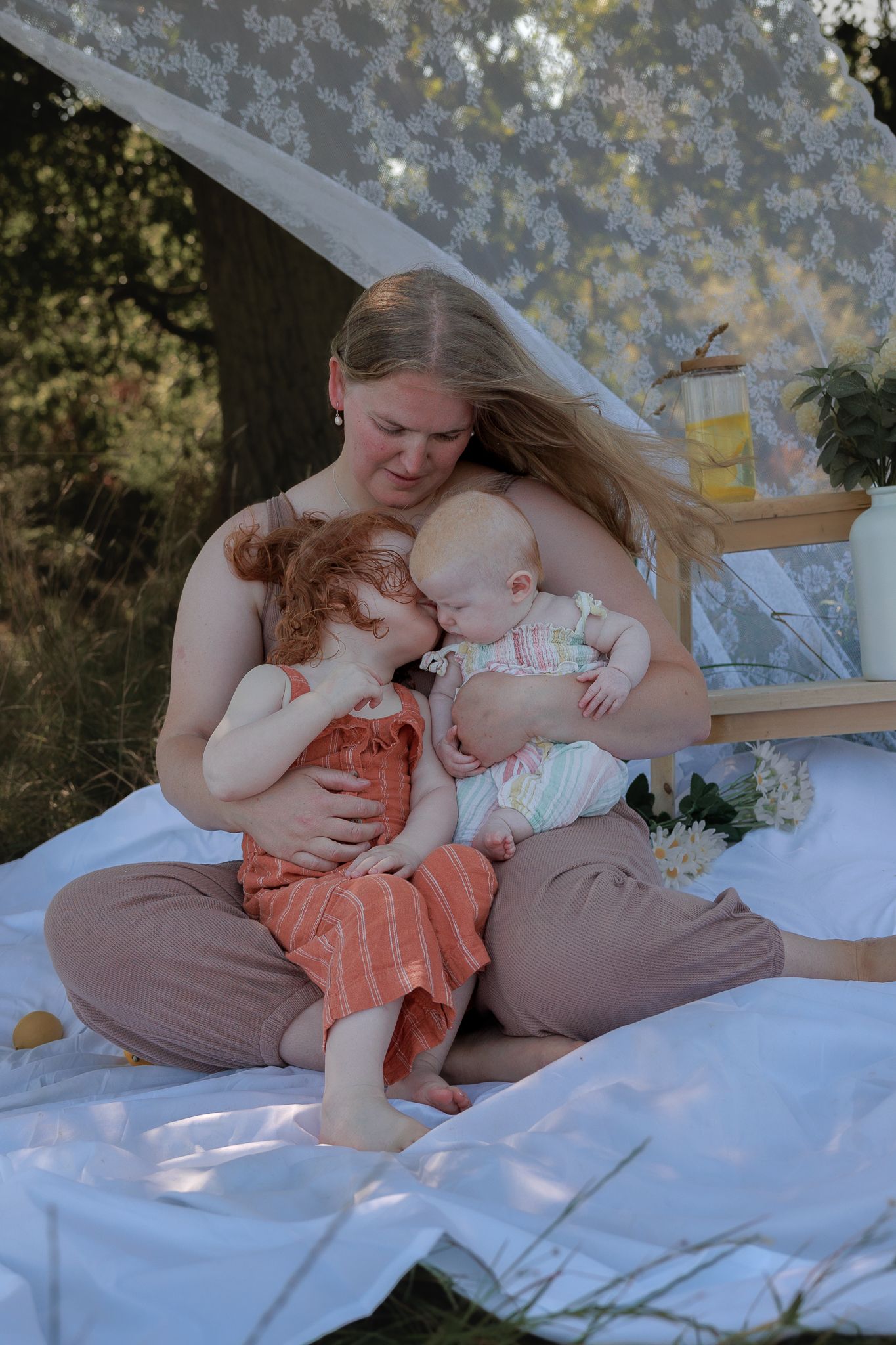 A mother sits outdoors on a white blanket, tenderly holding two young children under a floral canopy in Henley-on-Thames, Oxfordshire.
