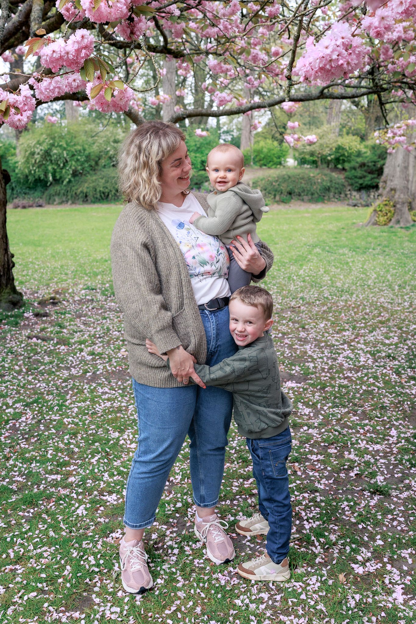 A smiling woman holds a baby while a young boy hugs her waist under blooming pink cherry blossom trees in a park in Chipping Norton.