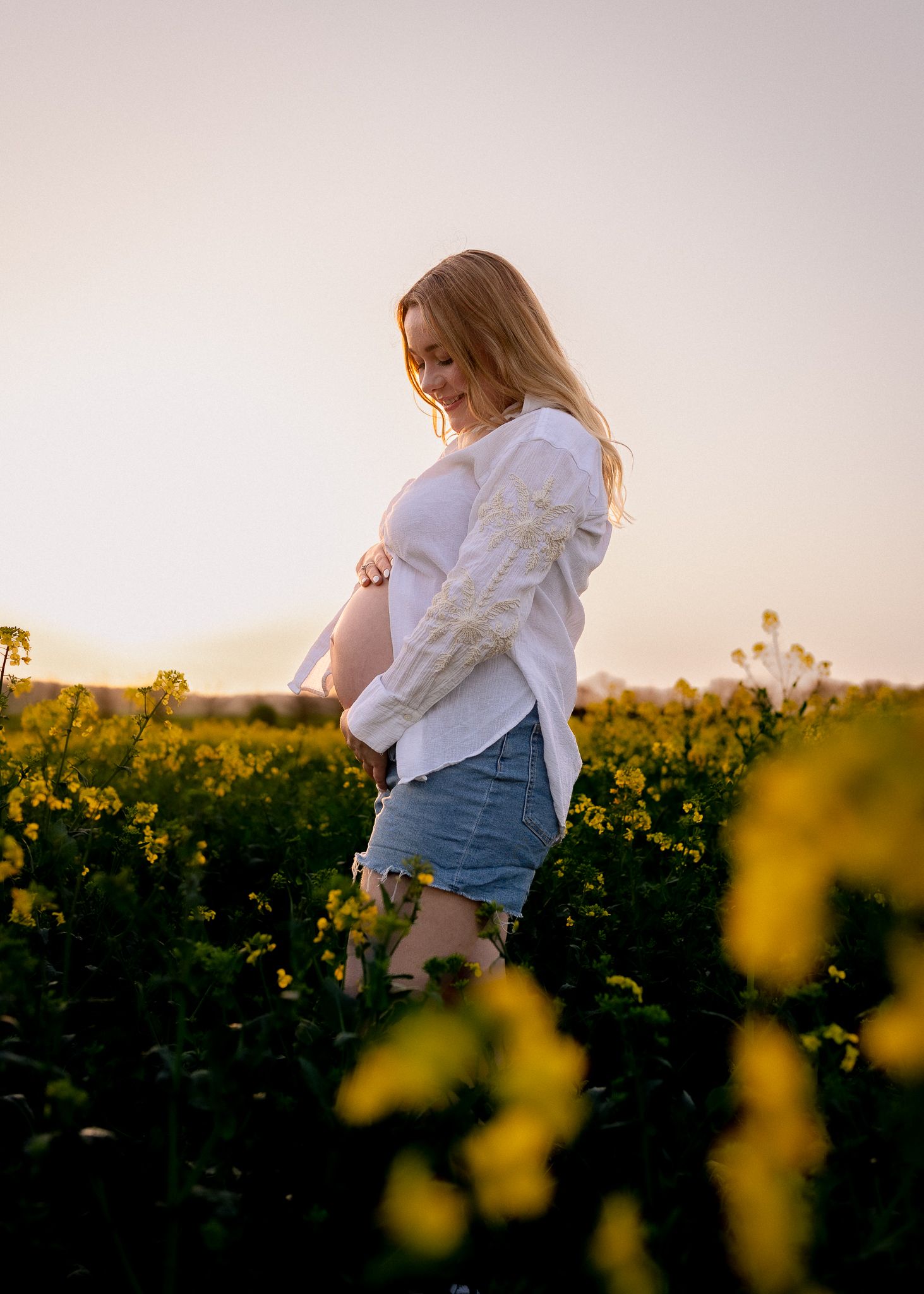 Pregnant woman in white shirt and denim shorts standing in yellow flower field at golden hour sunset in Marlborough, Wiltshire.