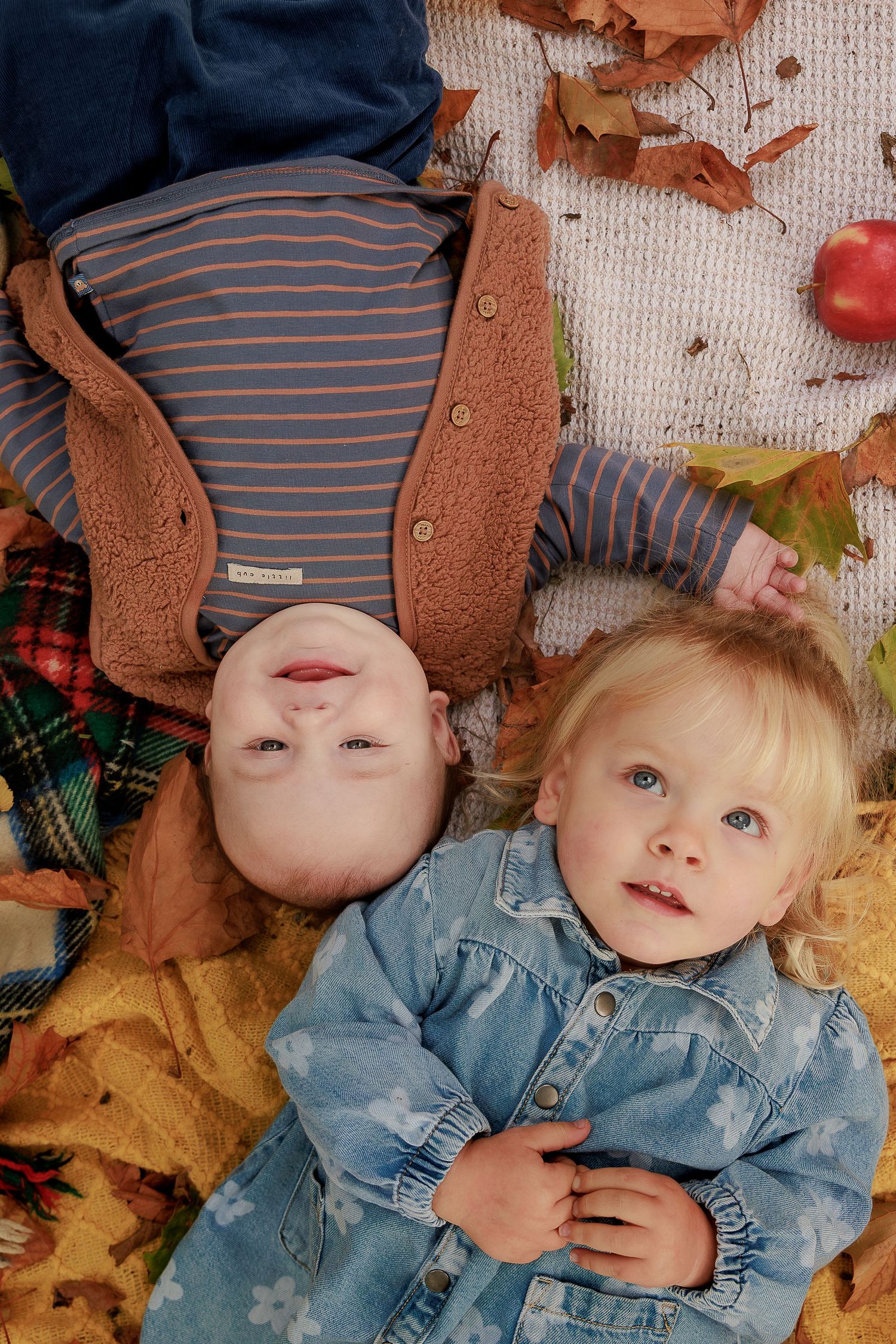 Two young children lying on a blanket surrounded by colorful autumn leaves, with a red apple nearby.