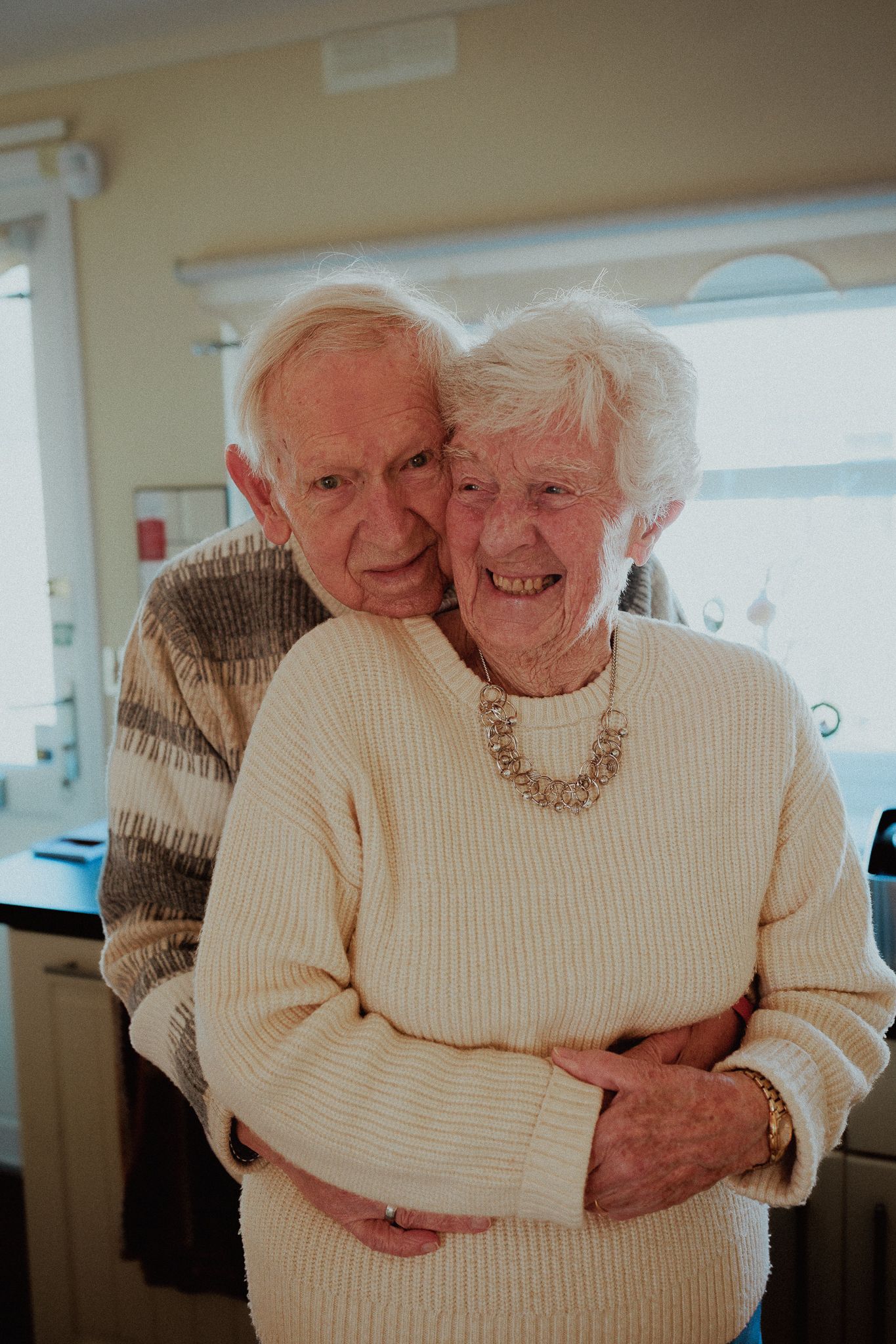 Elderly couple sharing a warm embrace, smiling together in a bright home interior in Bibury.