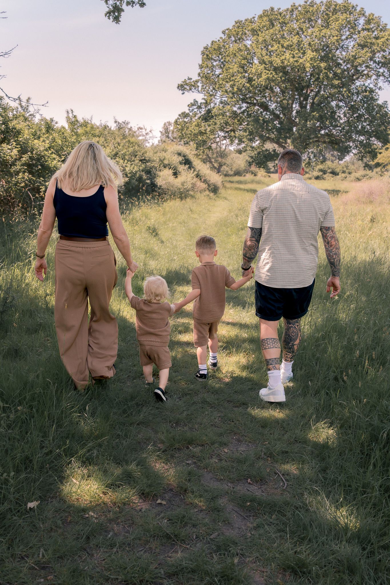 A family of four walks hand-in-hand through a lush green meadow, with two young children between tattooed dad and blonde mom.