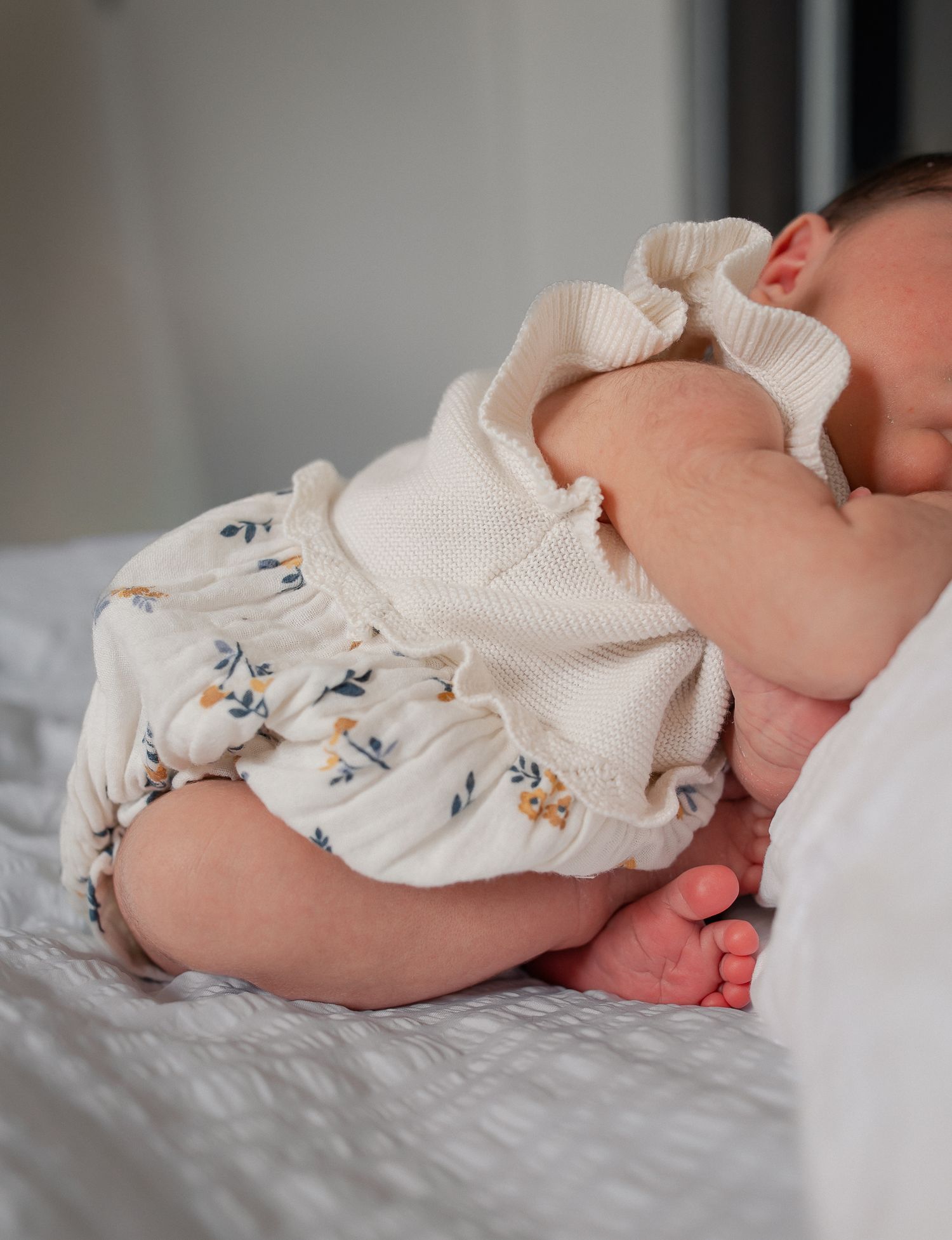 Newborn baby sleeping peacefully in a cream floral romper on a soft white bed.