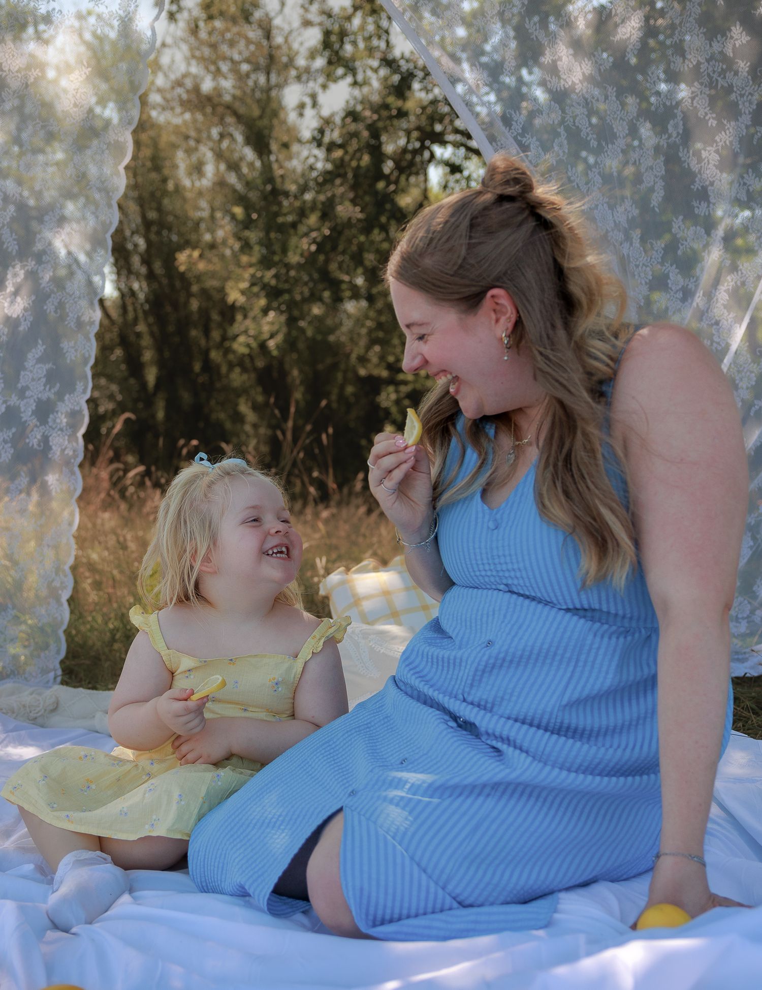 Pregnant woman in blue dress smiling with toddler girl in yellow dress during outdoor picnic photoshoot in Bibury, Cotswolds.