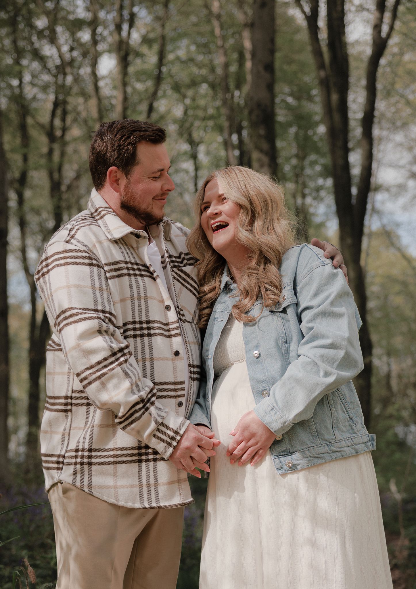 Smiling couple in a forest, woman in denim jacket and white dress showing baby bump, man in plaid shirt holding hands.