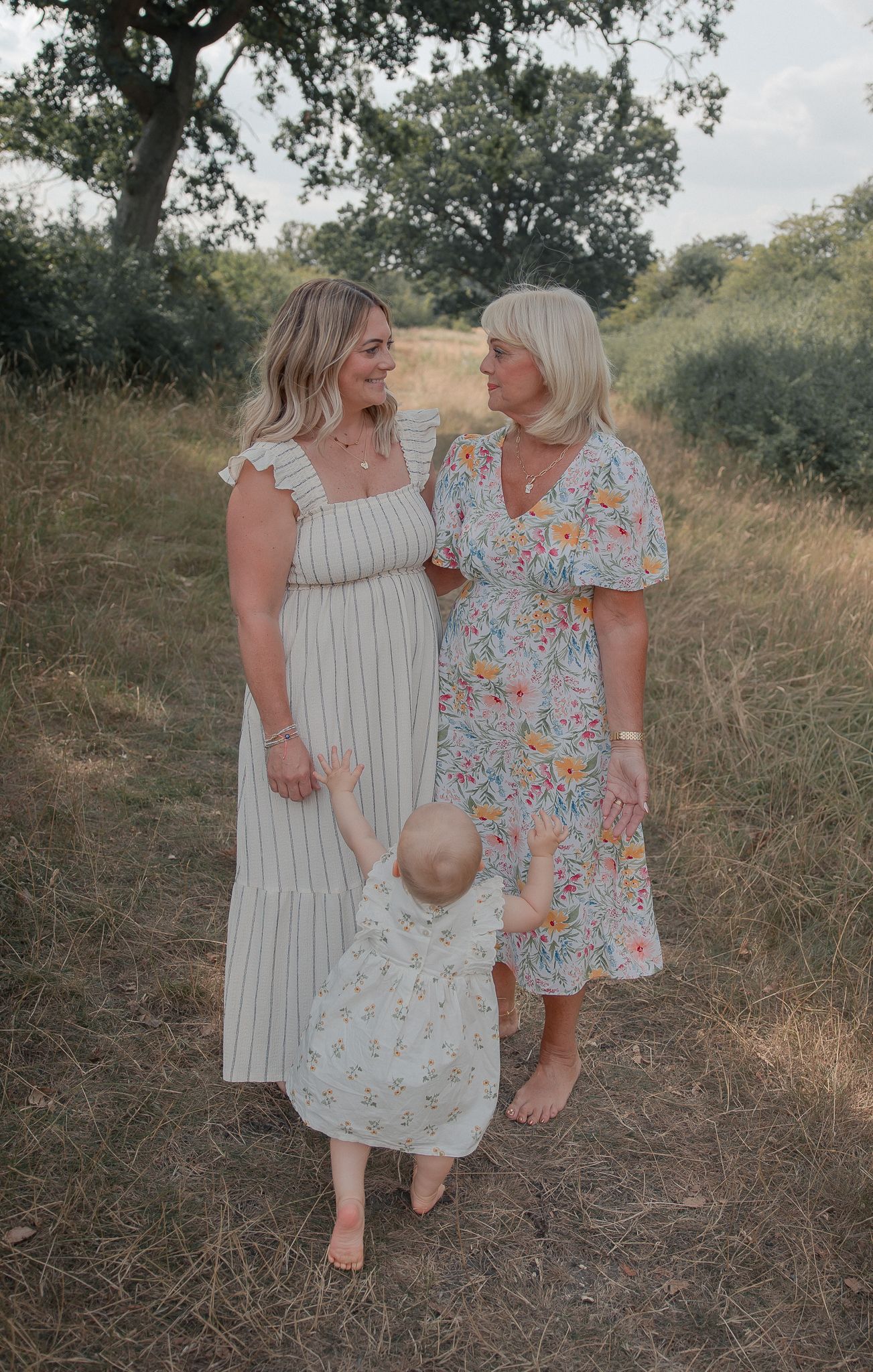 Two women in floral and striped dresses smile at each other while holding hands with a toddler on a sunny meadow path.
