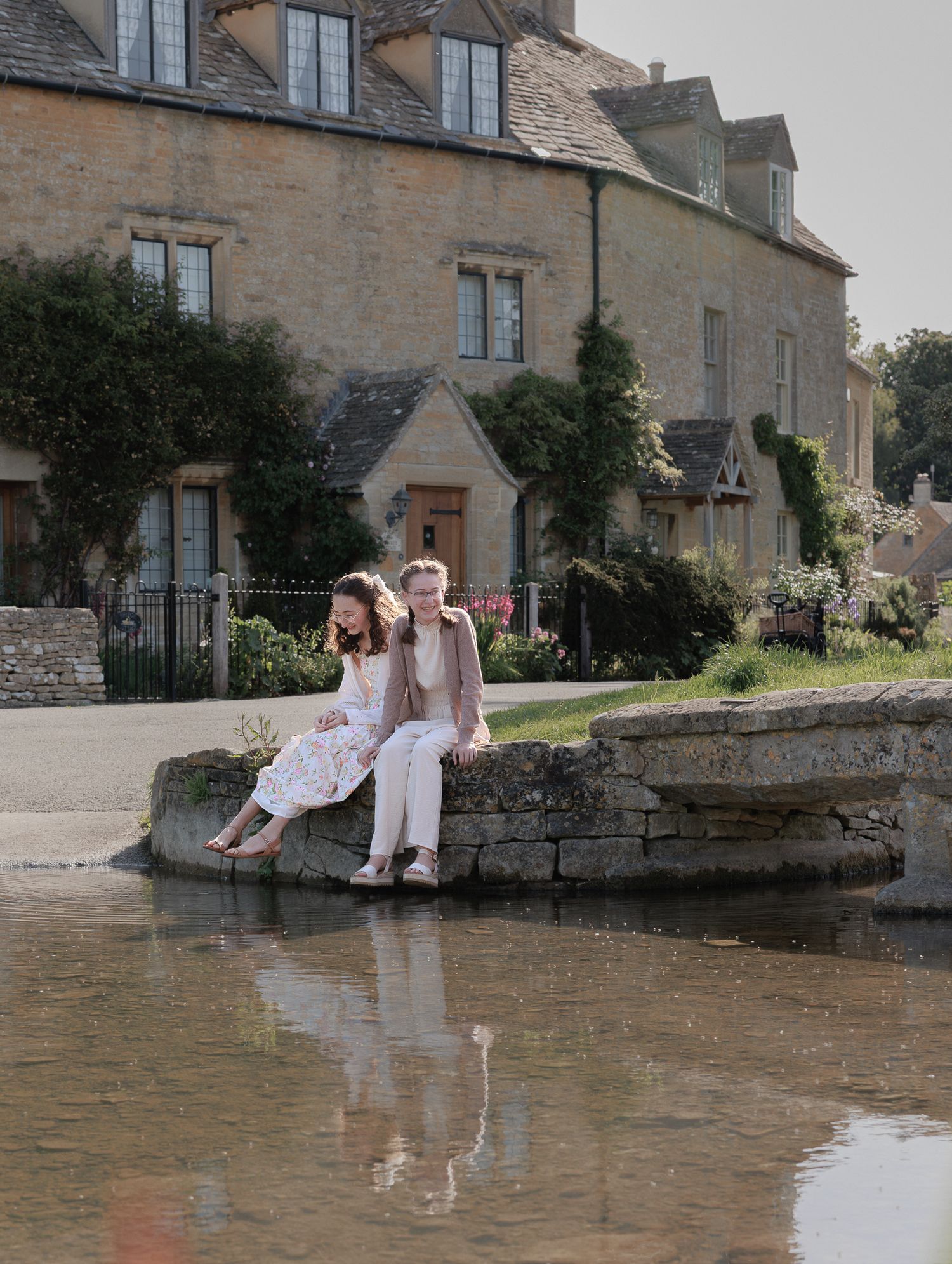 Two people sitting on a stone wall by a reflective pond near a charming Cotswolds stone cottage on a sunny day near Bourton-On-The-Water.