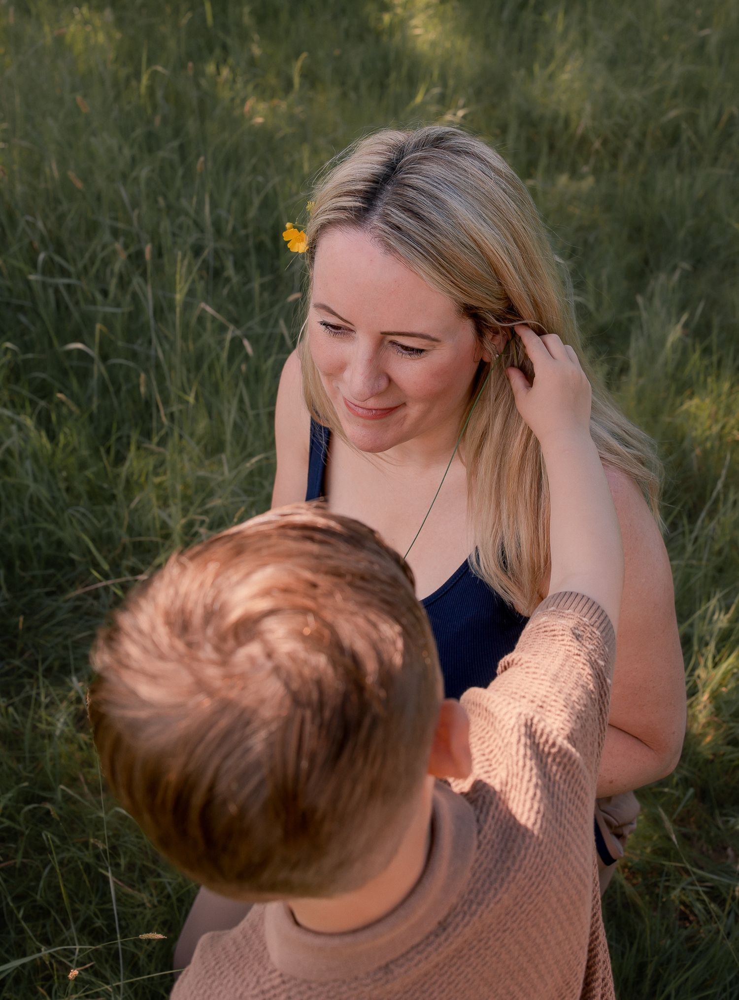 A blonde woman smiles as a child places a yellow flower in her hair in a sunny green field.