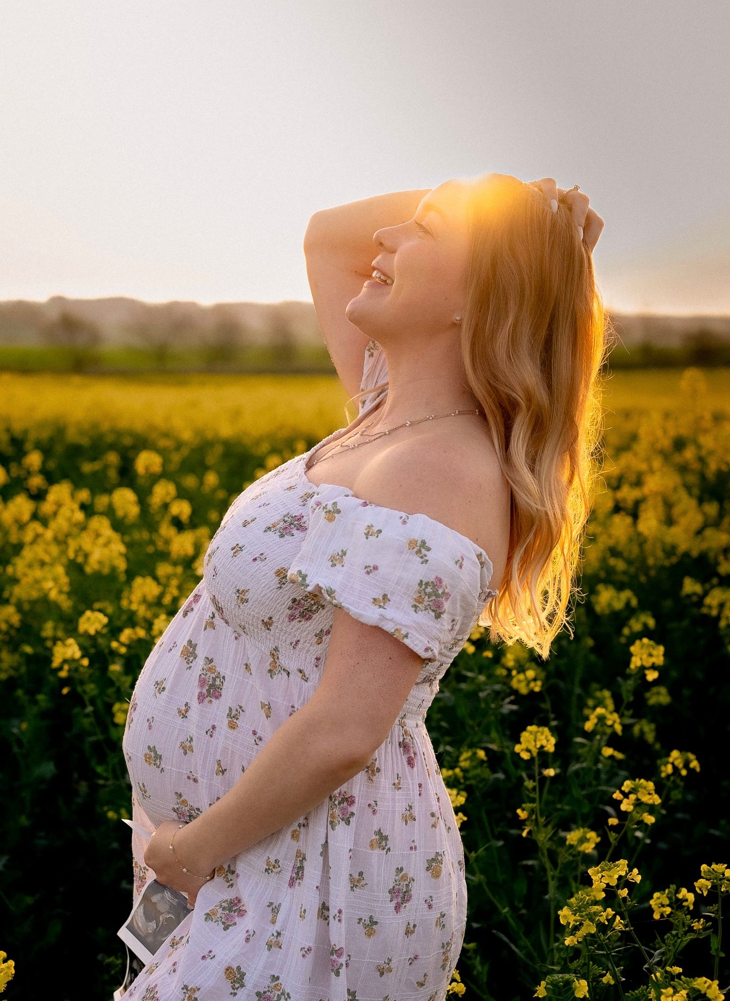 Pregnant woman in floral dress standing in yellow canola flower field at golden sunset in Marlborough.