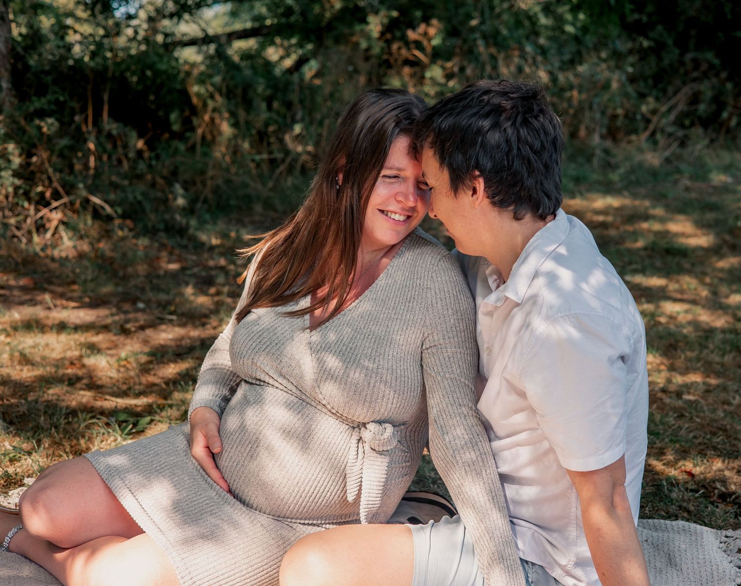 Pregnant woman and partner sitting together outdoors on a blanket, sharing a tender moment in Weybridge.