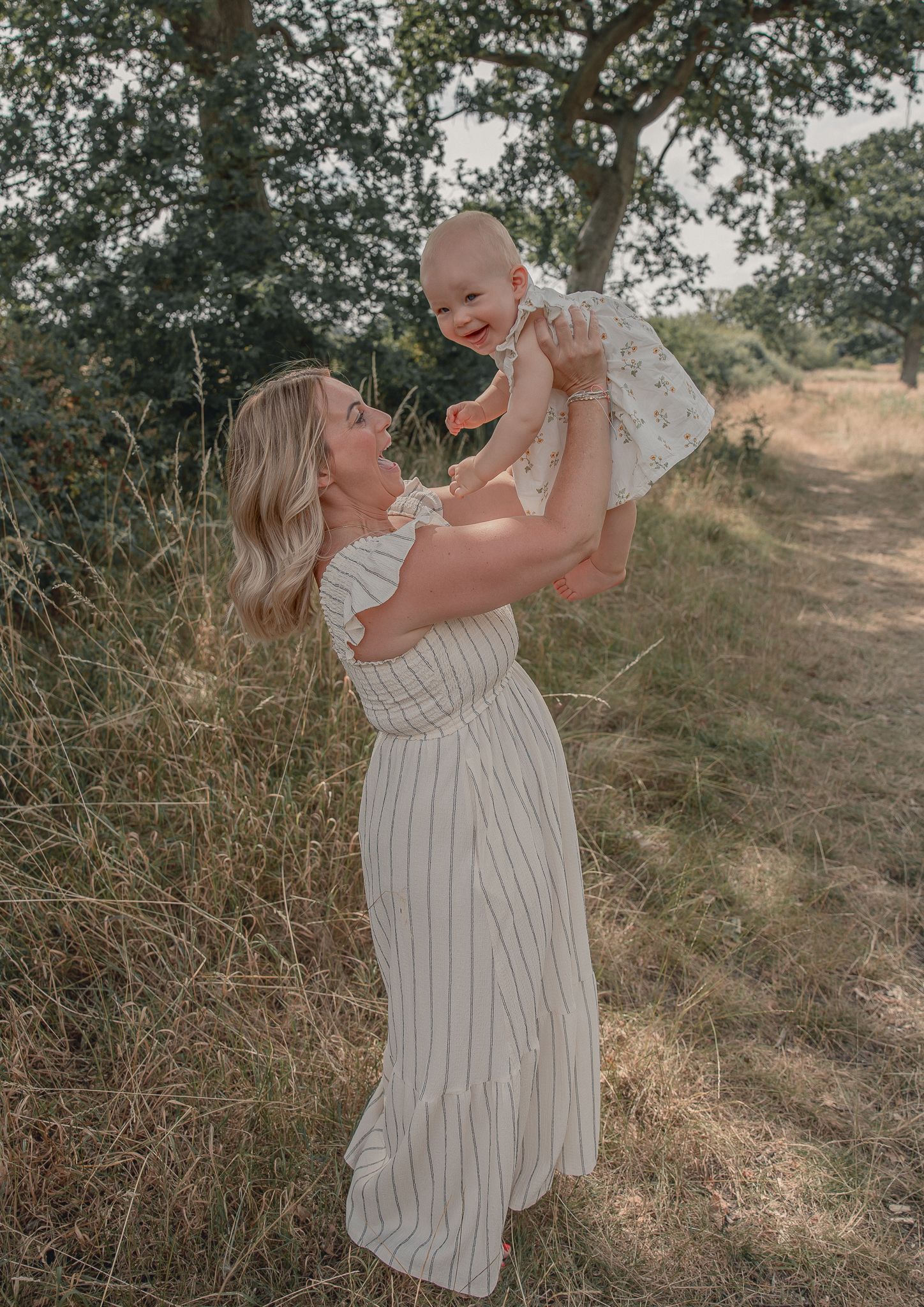 A woman in a striped maxi dress lifts a smiling baby girl in a sunny outdoor field with trees.