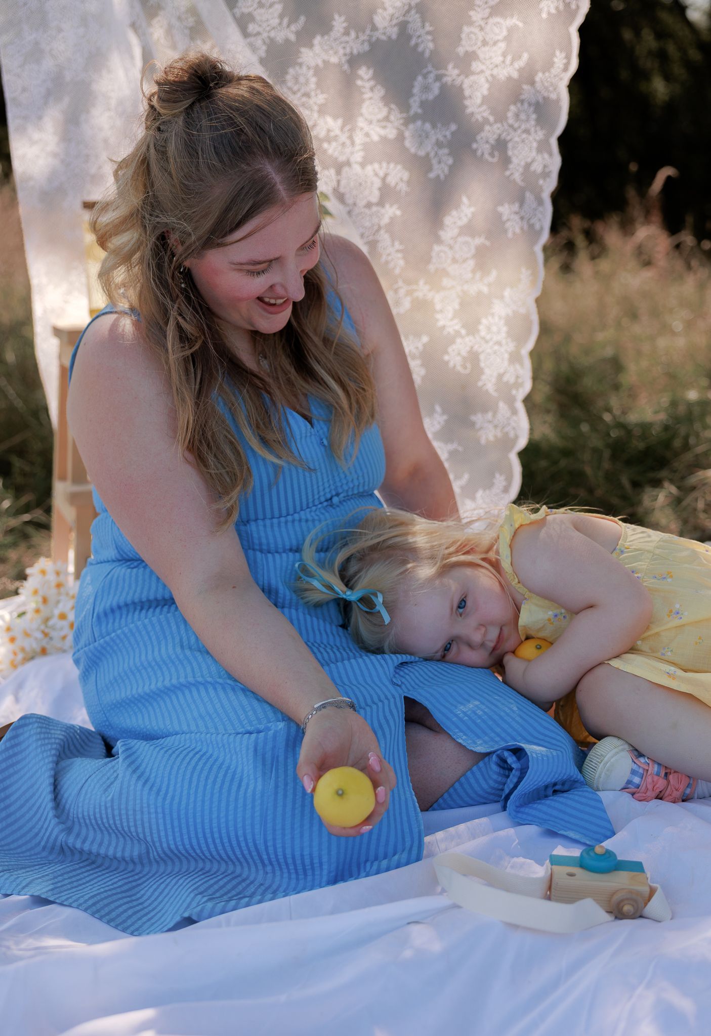 A woman in a blue dress sits on a blanket outdoors, smiling at a young girl in a yellow dress in Bibury, Cotswolds.
