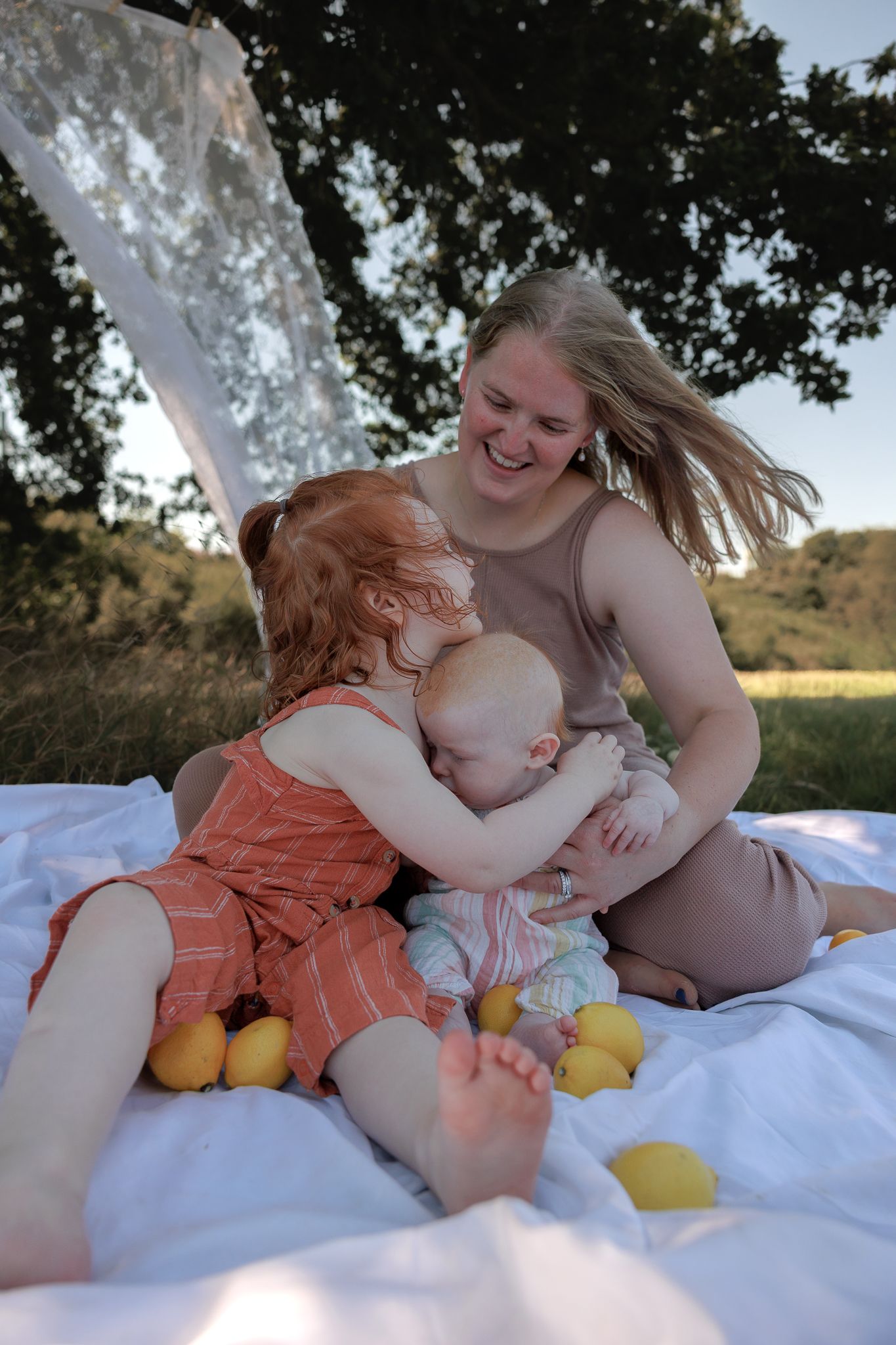 A smiling mother sits on a white blanket outdoors while her redhead toddler hugs a baby, with lemons scattered around in Summertown, Oxford.