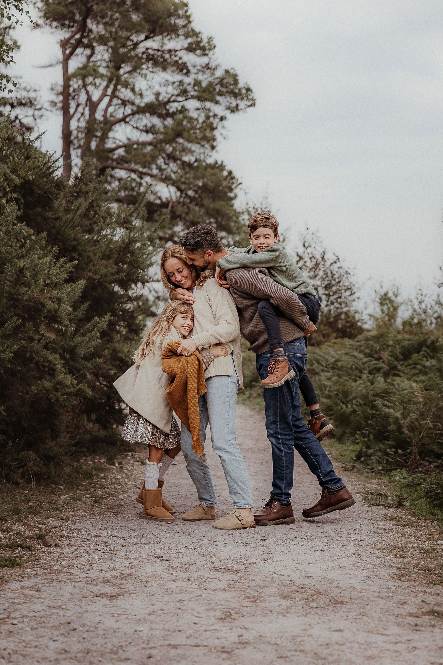 Family of four embracing in a group hug on a sandy forest path, with a boy lifted onto his dad's back and a girl in a mustard scarf.