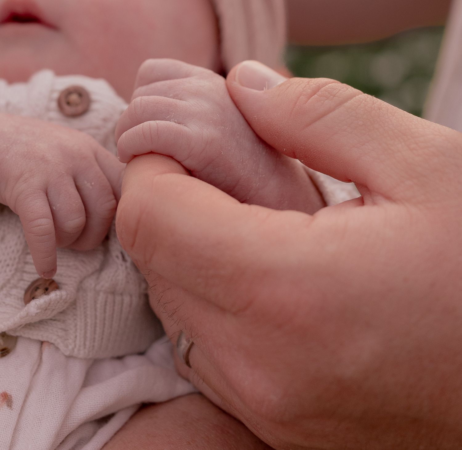 A newborn baby grasping an adult finger with tiny delicate hands, wearing a soft pink knit outfit.