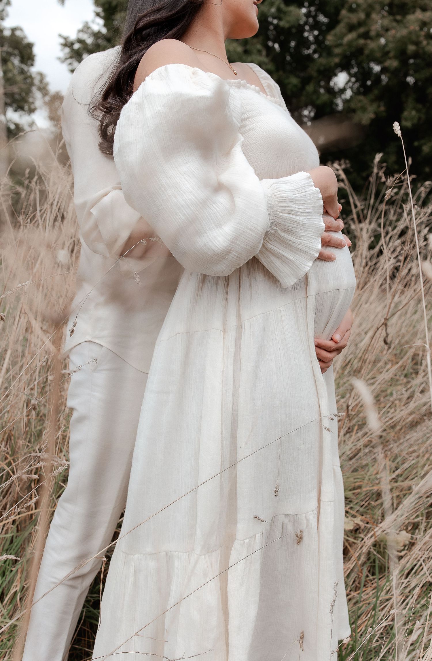 Pregnant woman in white off-shoulder dress with ruffled sleeves standing in golden field grass during maternity photoshoot.