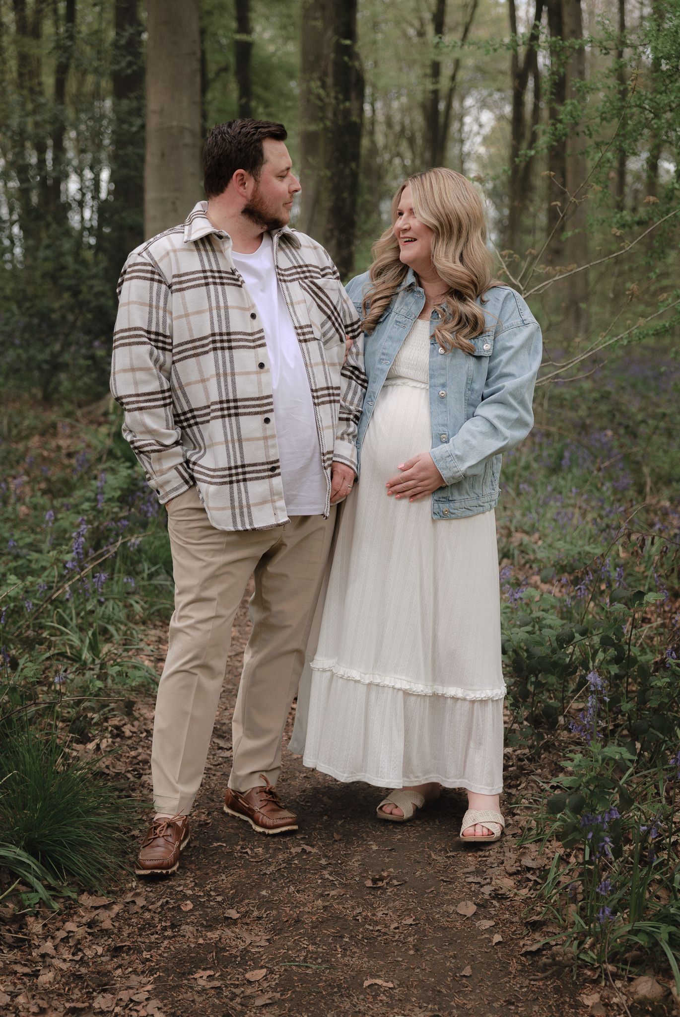 Pregnant woman in white dress and denim jacket with partner on woodland path surrounded by bluebells.