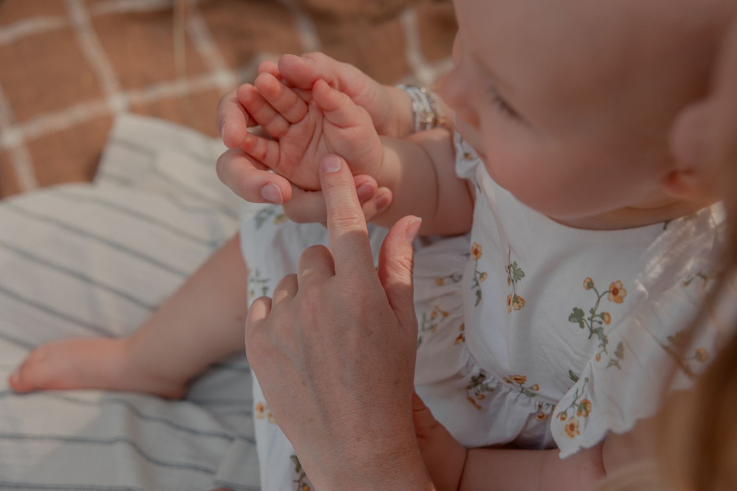 A baby in a floral outfit gently chews on an adult's finger while being held in a warm, cozy setting.