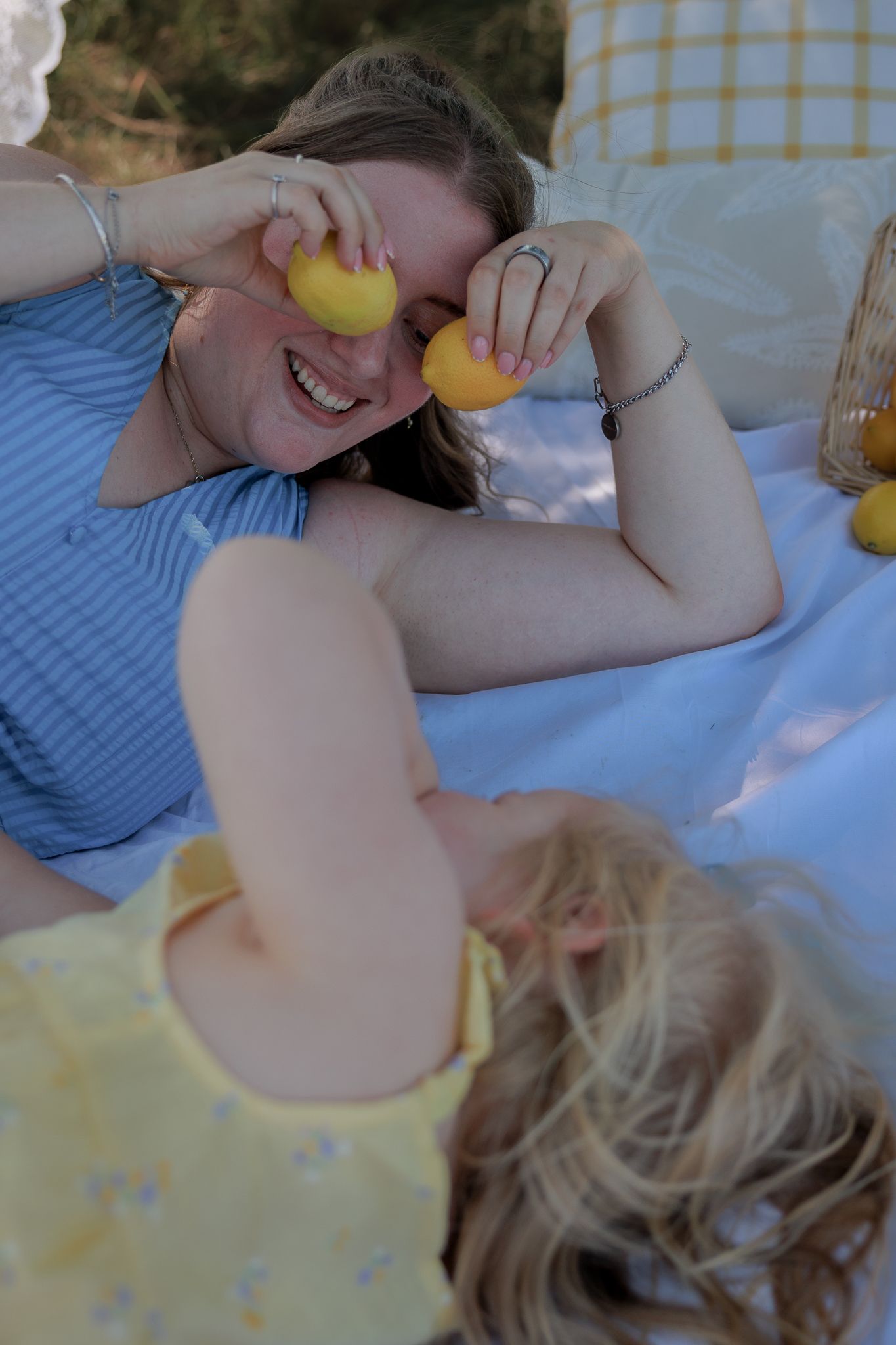 Woman and child laughing at a picnic, holding lemons over their eyes while lying on a blue blanket outdoors in Bibury, Cotswolds.