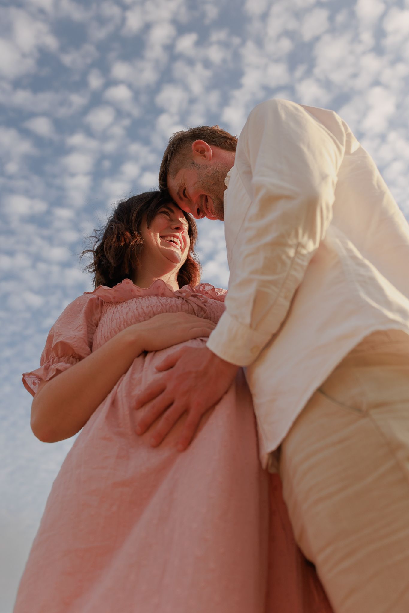 A smiling couple in soft pink and white linen outfits embrace outdoors under a cloudy blue sky for a Maternity Photoshoot, Southstoke.