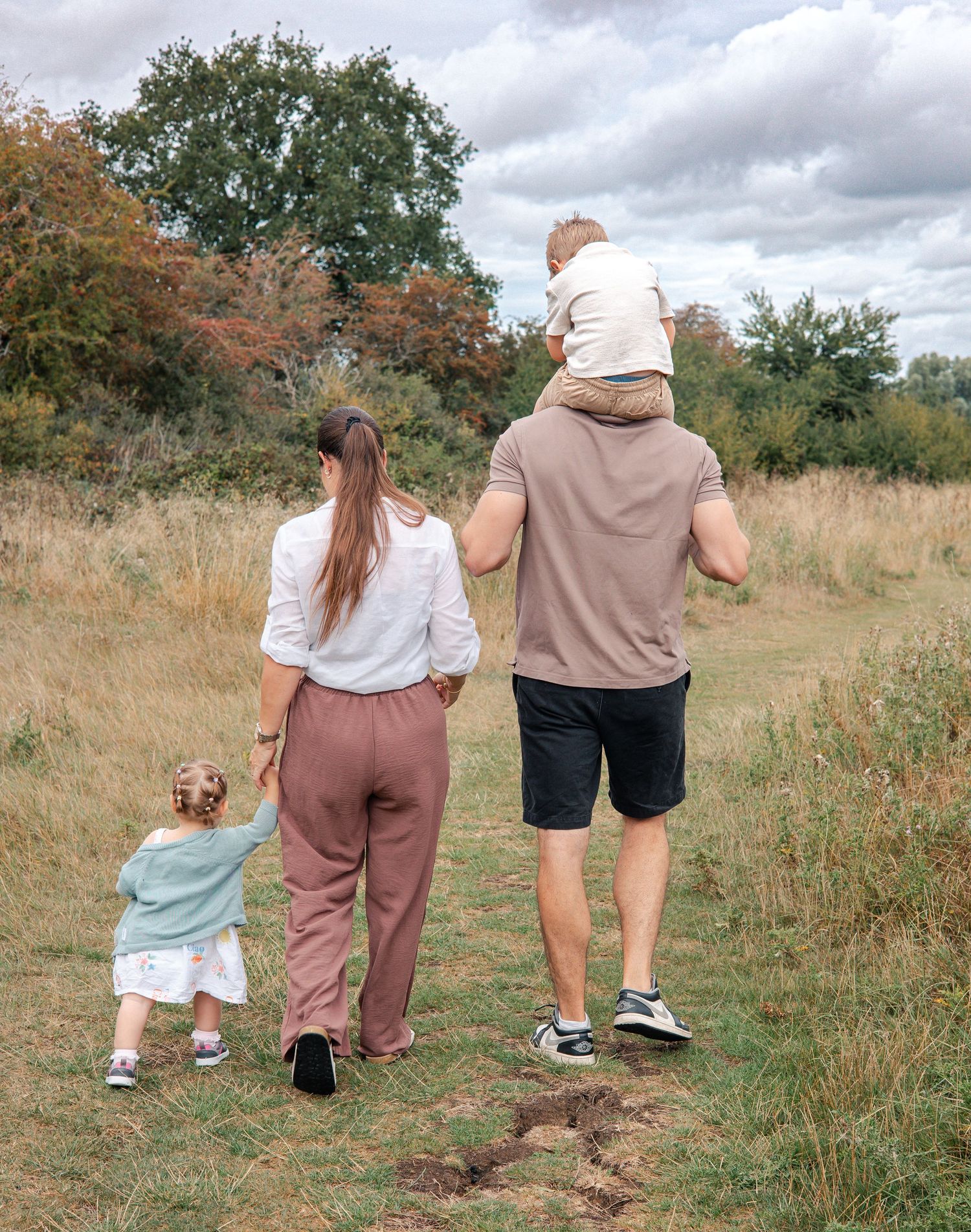 Family walking together on a dirt path through an autumn meadow with trees and cloudy skies in the background. Carterton.