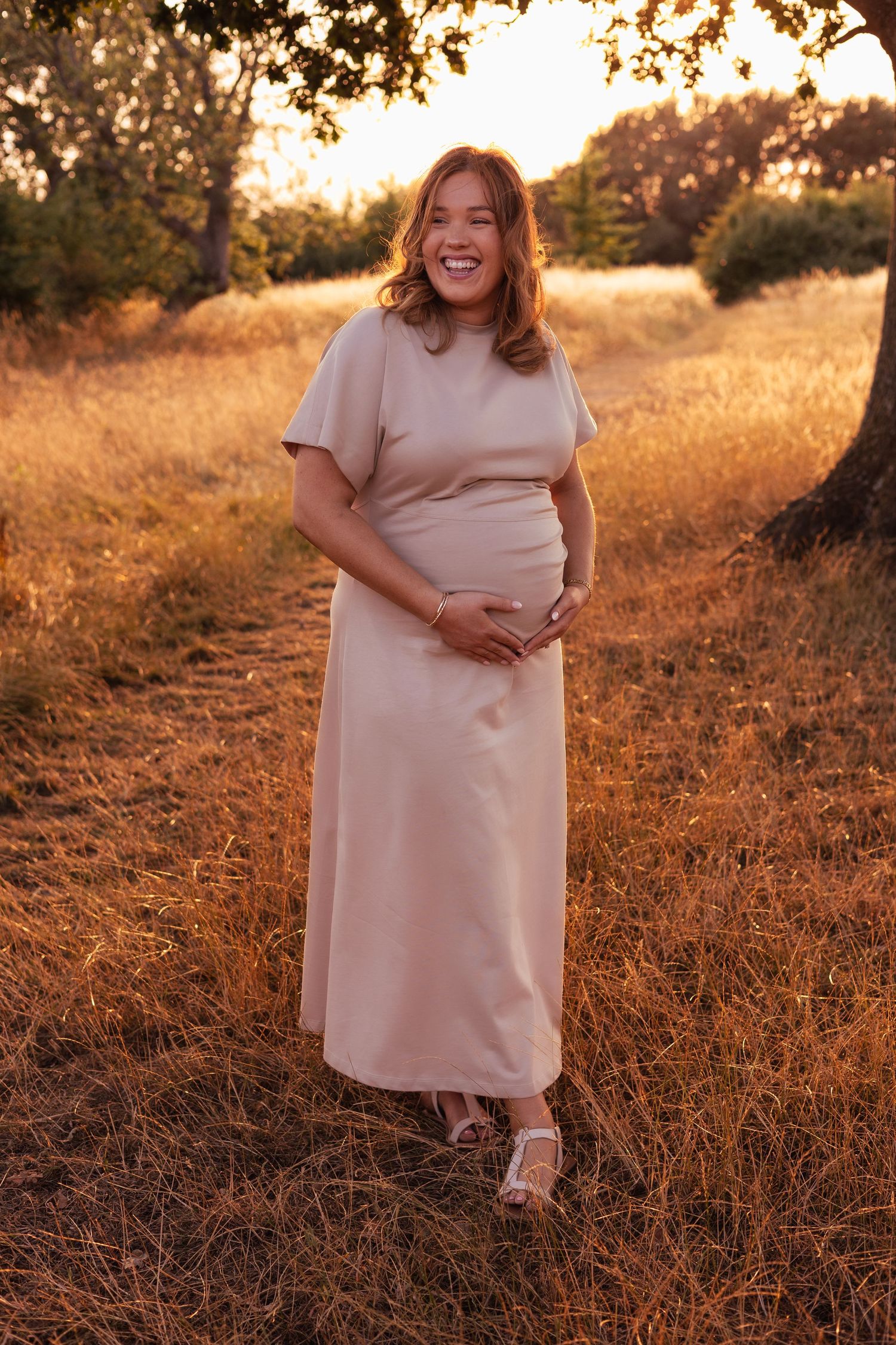 A pregnant woman in a pink dress stands in a field on a sunny day.