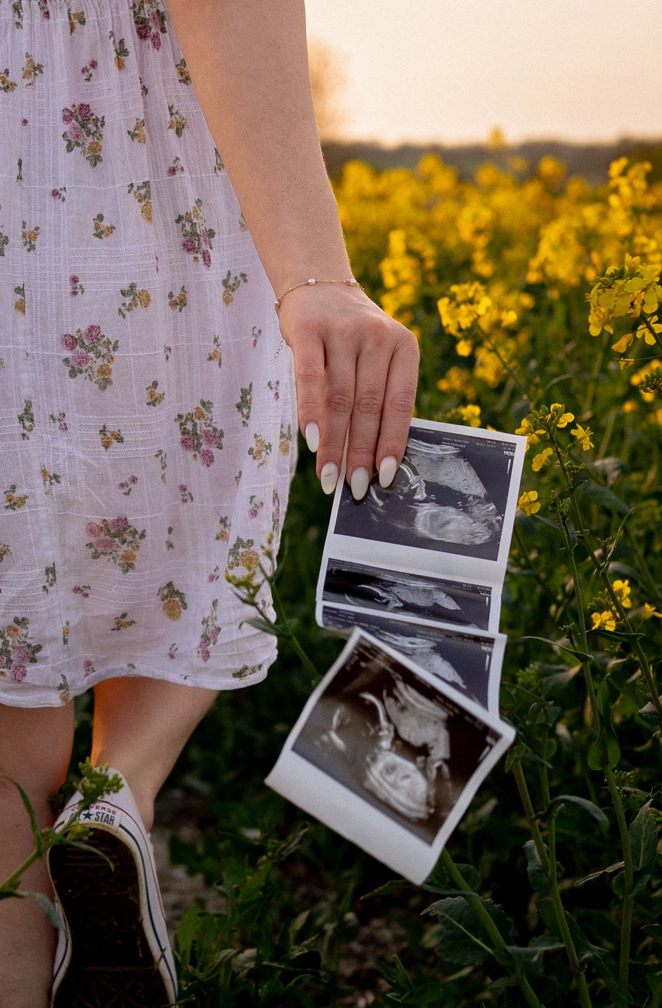 Pregnant woman in floral dress holding ultrasound photos in a yellow rapeseed flower field at sunset in Abingdon, Oxford.