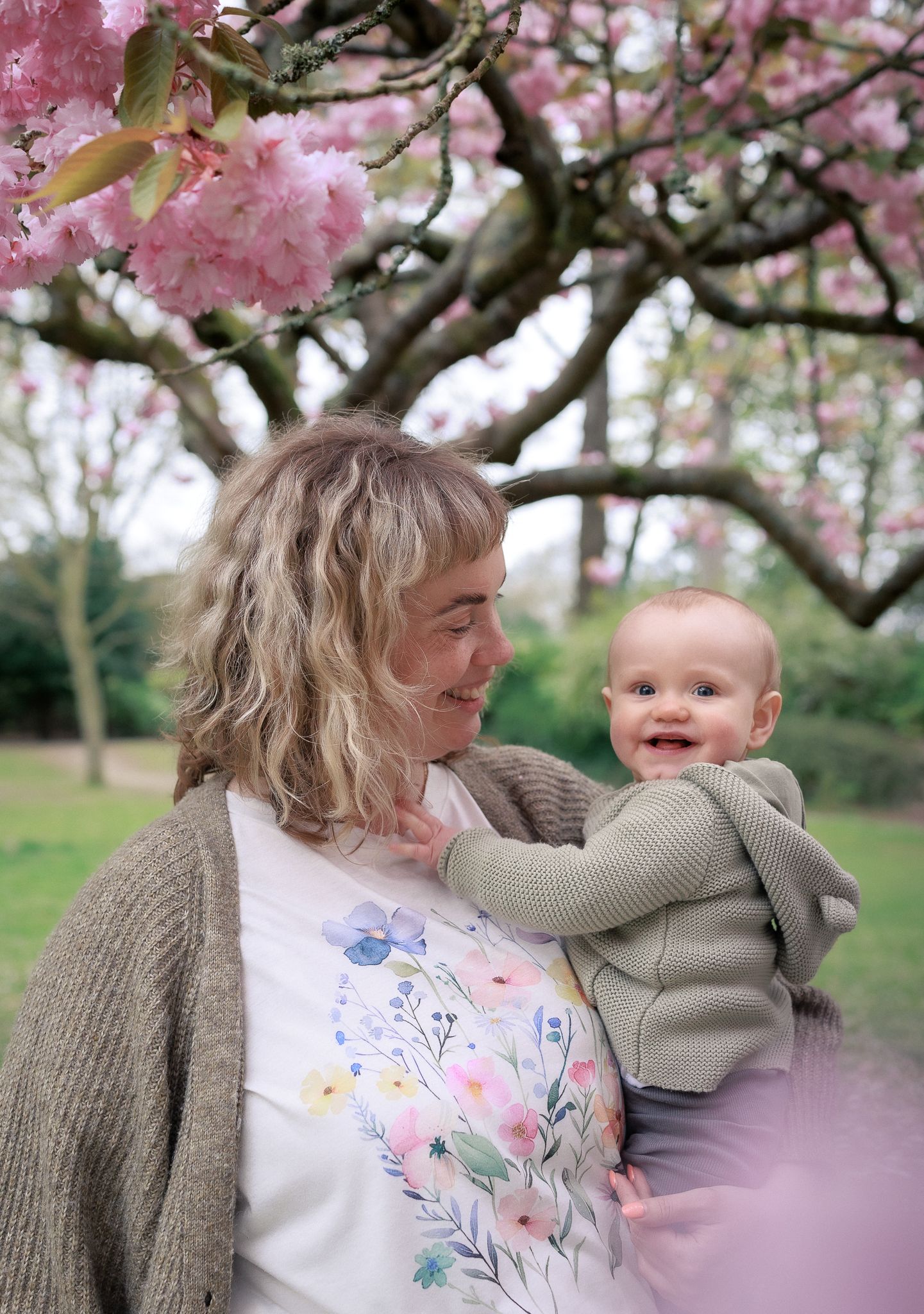 A smiling mother holds her happy baby under a blooming pink cherry blossom tree in spring in Bourton-on-Water.
